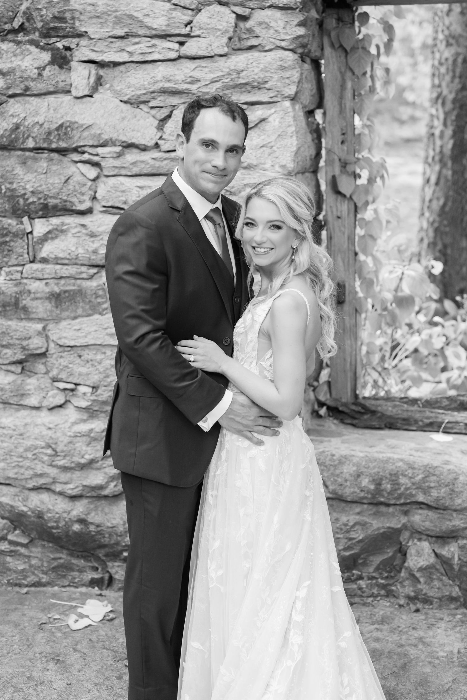 Black and white romantic wedding portraits of couple against rustic stone wall in elegant attire.