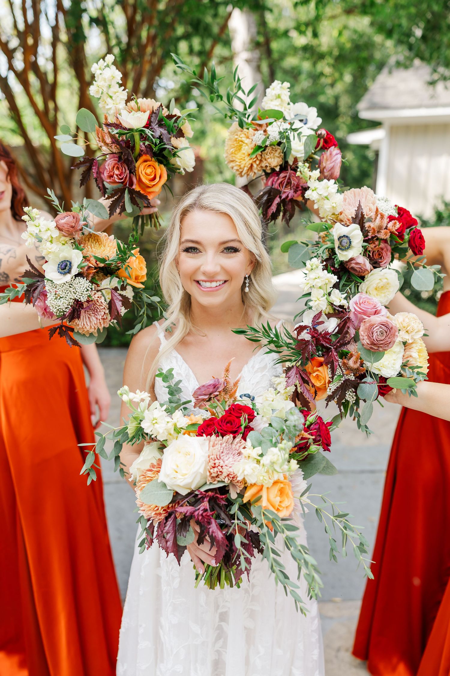 Bridal party in burnt orange dresses holding vibrant autumn-colored bouquets with roses and mixed florals at outdoor wedding.
