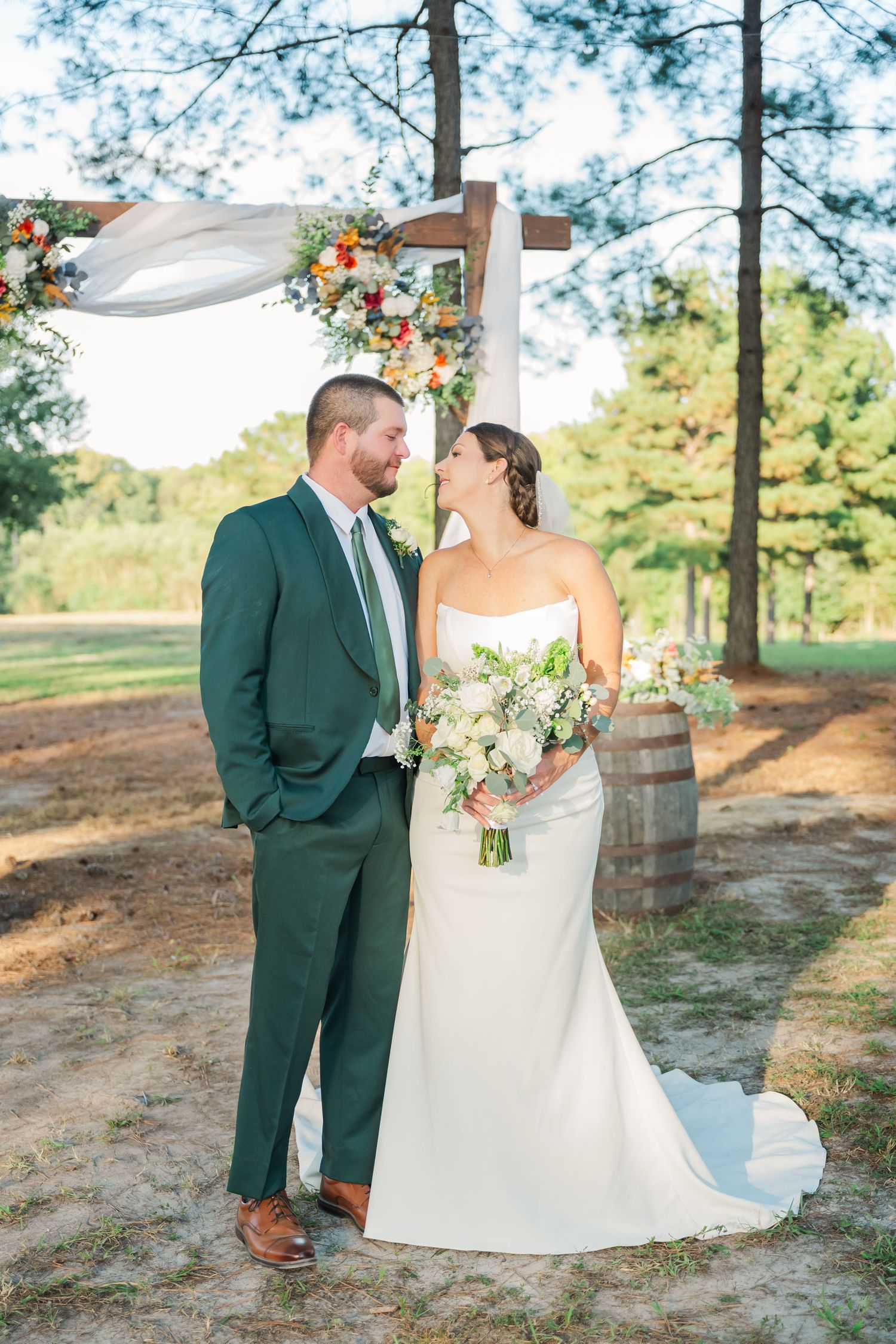 Bride and groom pose under a floral wooden arch outdoors, she in a white gown holding a bouquet, he in a dark green suit.