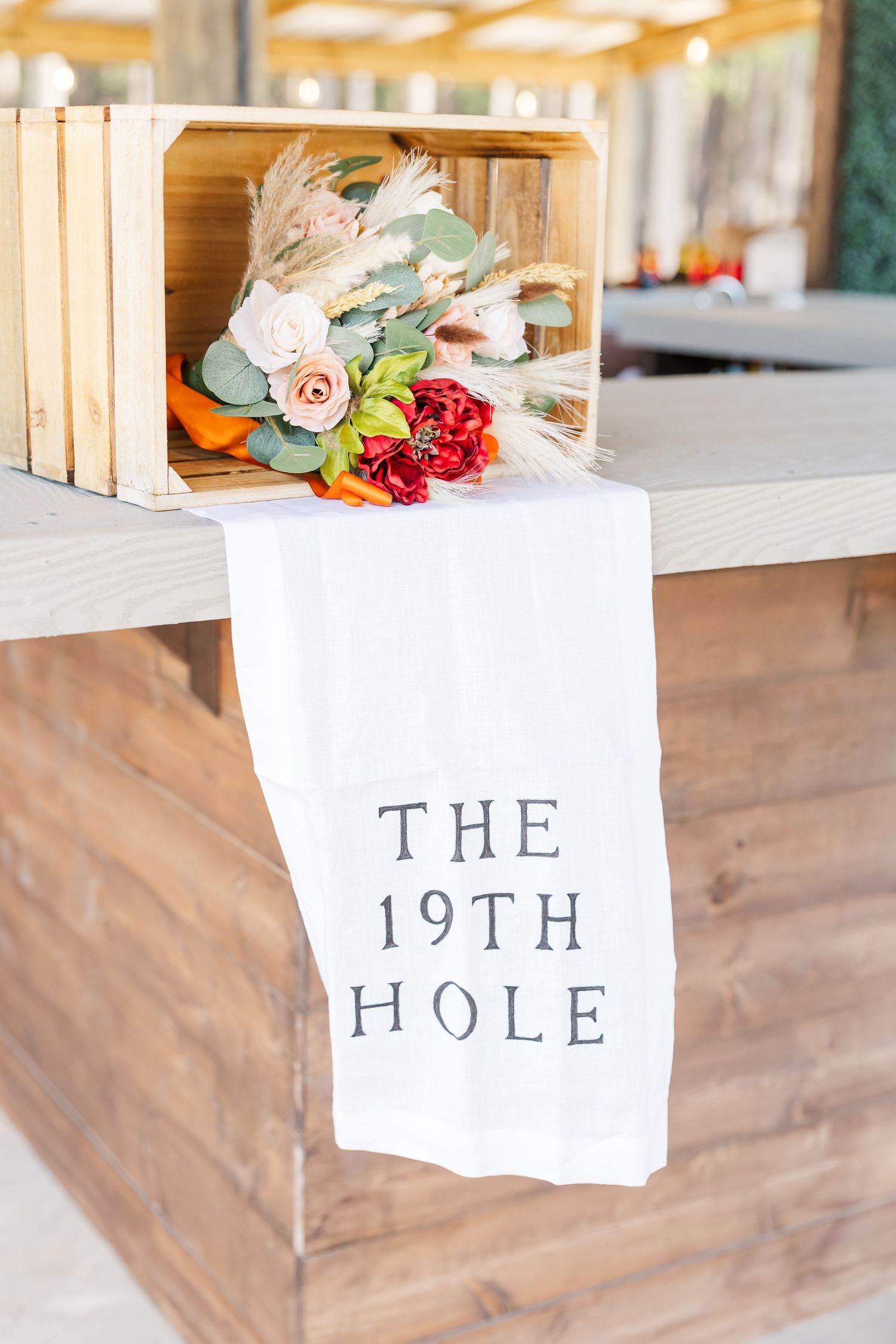 White towel reading The 19th Hole draped over a wooden bar, with a floral bouquet in a rustic crate.