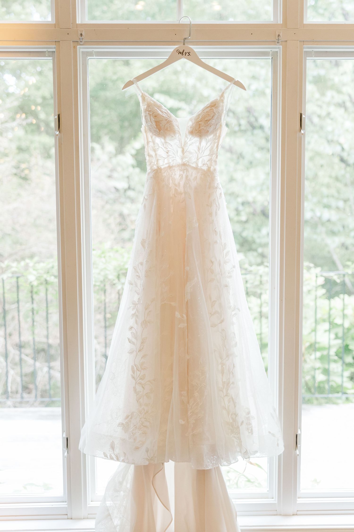 Three elegant white wedding dresses hanging on wooden hangers against bright windows in a bridal suite.
