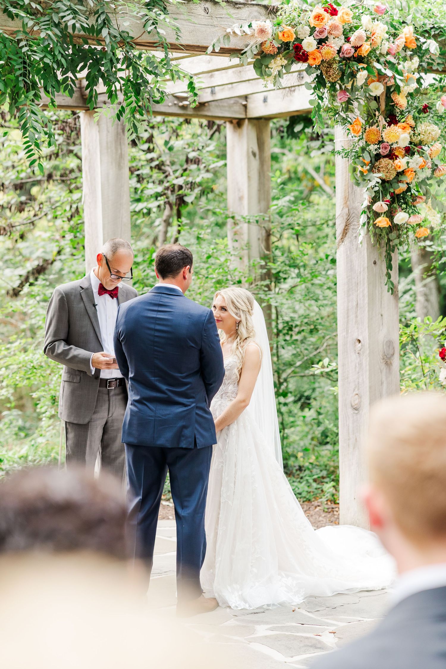 Outdoor wedding ceremony under wooden pergola decorated with lush orange and peach floral arrangements.