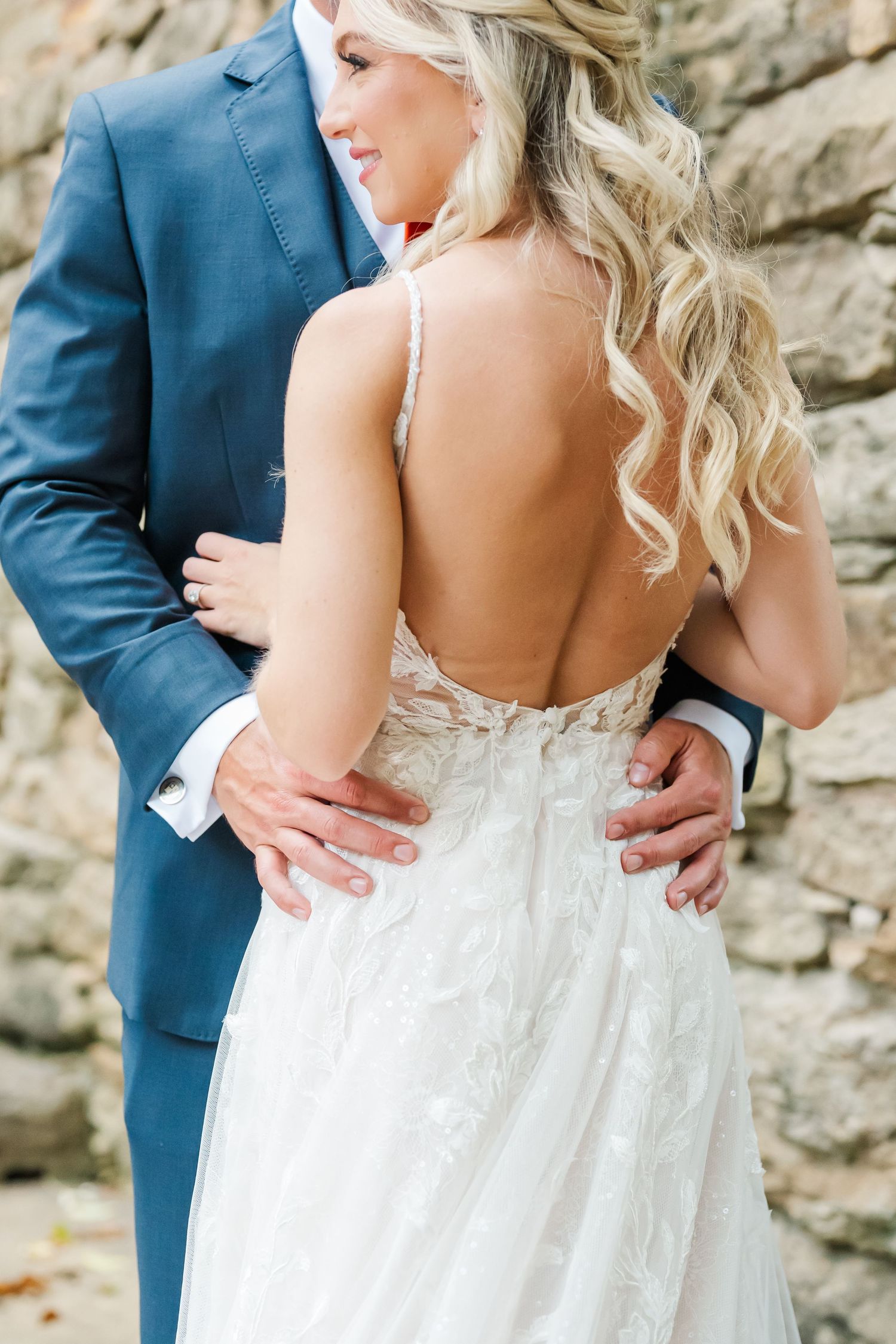 A close-up view of a wedding couple embracing, showing the bride's backless lace gown and groom's navy suit.
