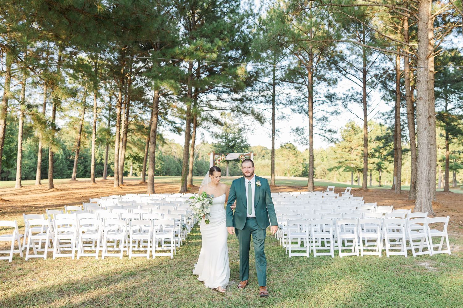 Outdoor wedding ceremony setup with white chairs arranged in rows among tall pine trees on a sunny day.
