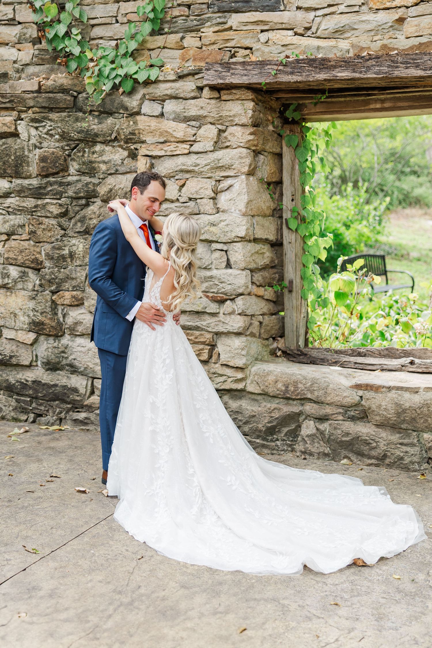 A romantic wedding couple embraces against a rustic stone wall with trailing vines in an intimate outdoor setting.