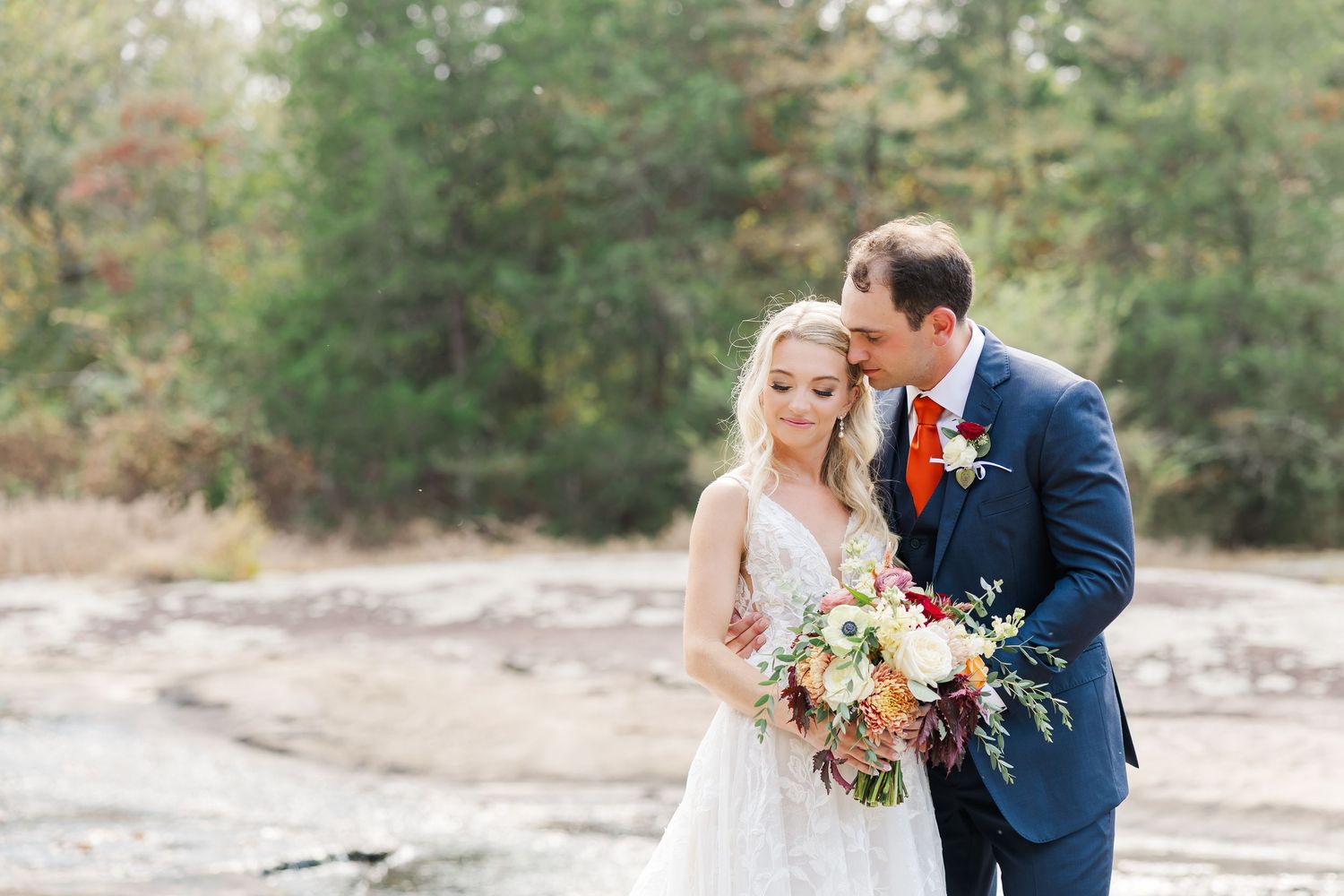A couple in formal attire walks together on a rocky beach path surrounded by autumn foliage.