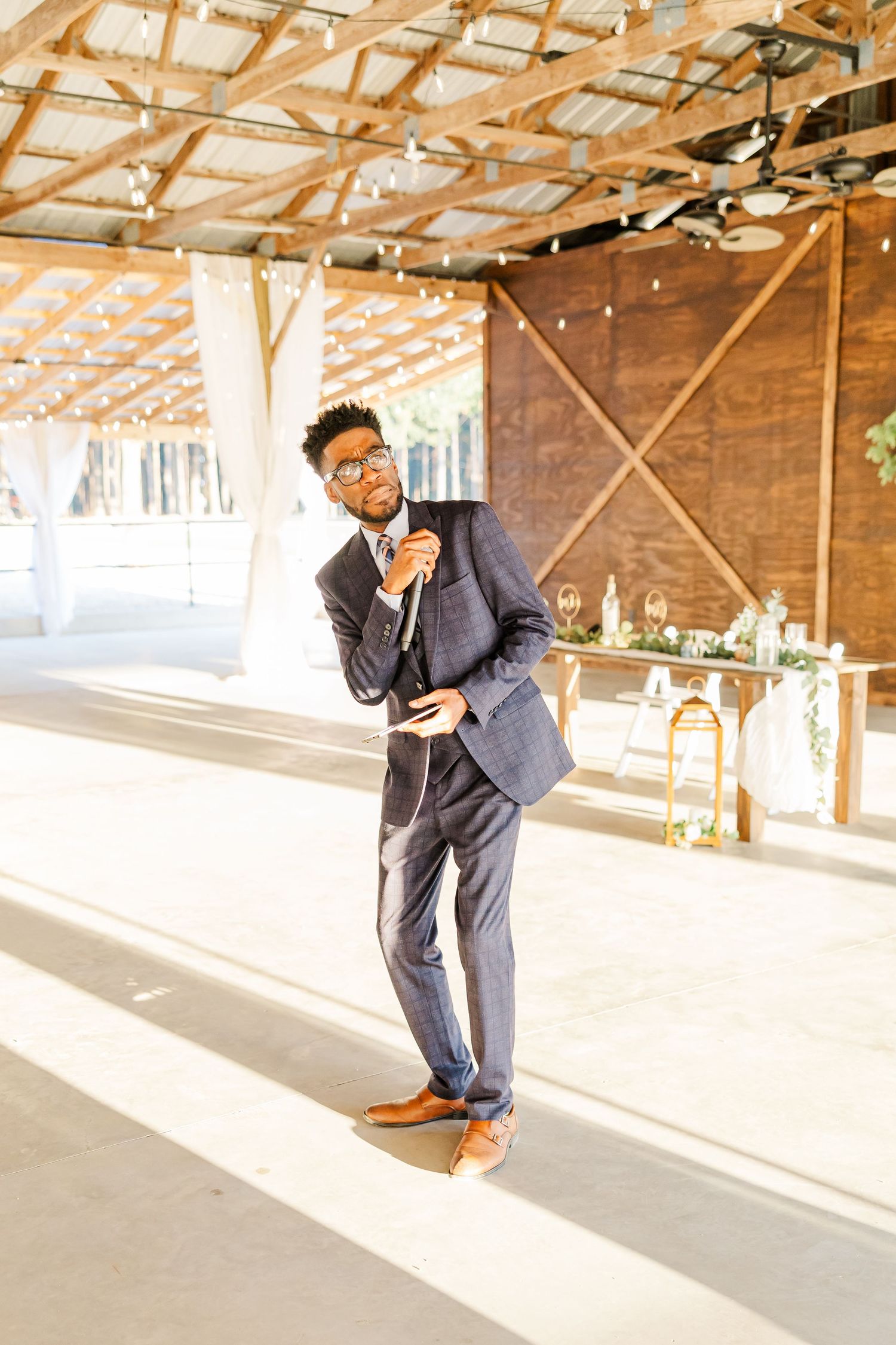 Stylish groom in navy suit and brown shoes posing confidently at rustic barn wedding venue.