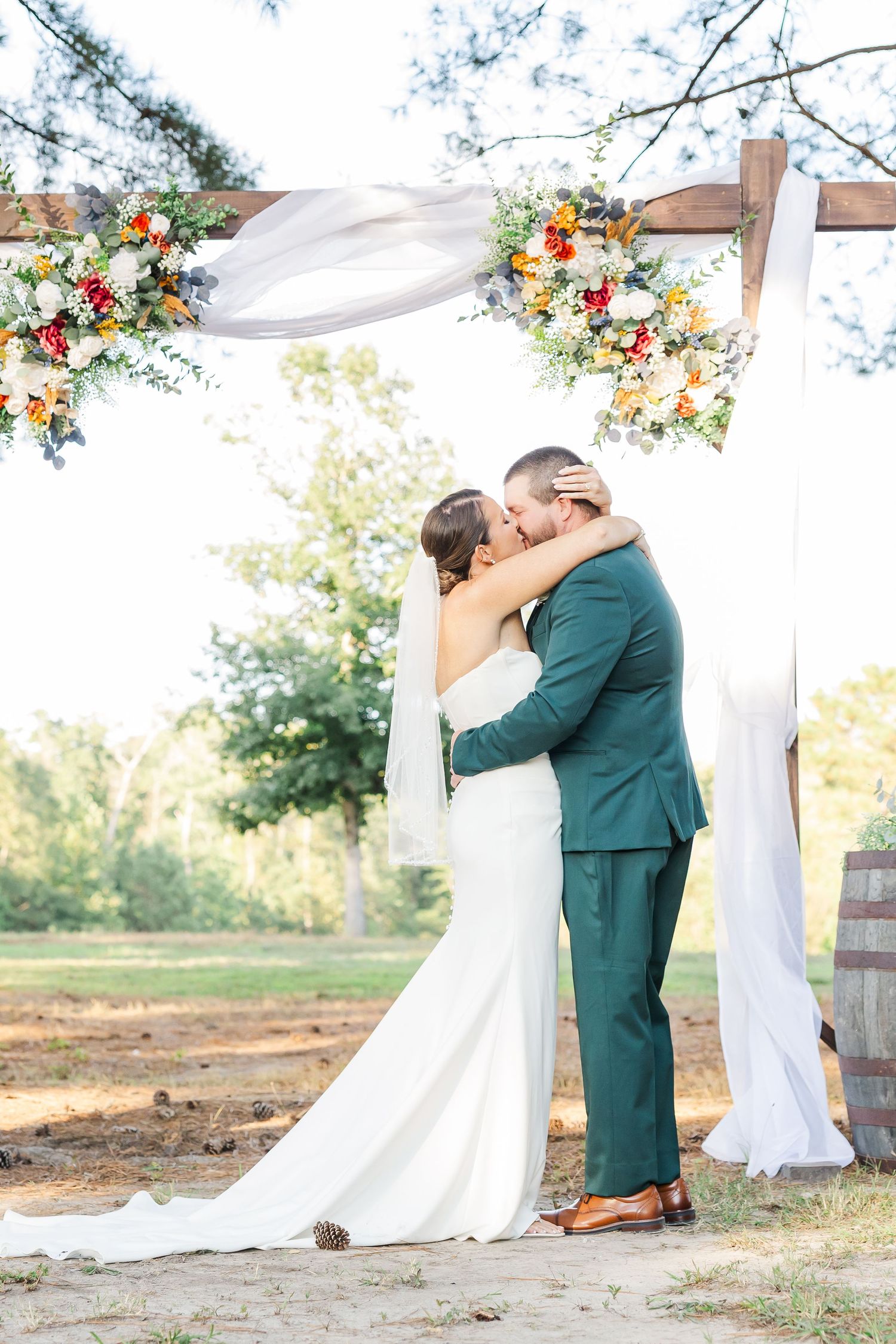 Bride and groom share first kiss under a floral wooden arch with white draping at an outdoor fall wedding ceremony.