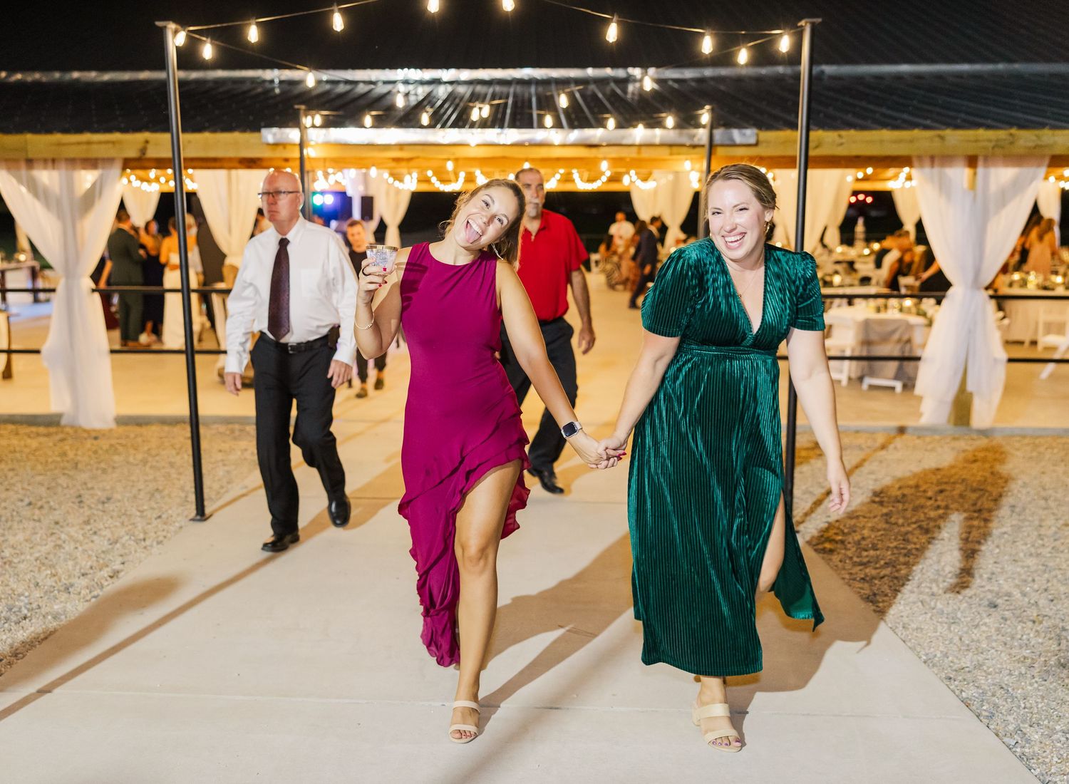 Two women in elegant magenta and green gowns walk joyfully at an outdoor wedding reception under string lights.
