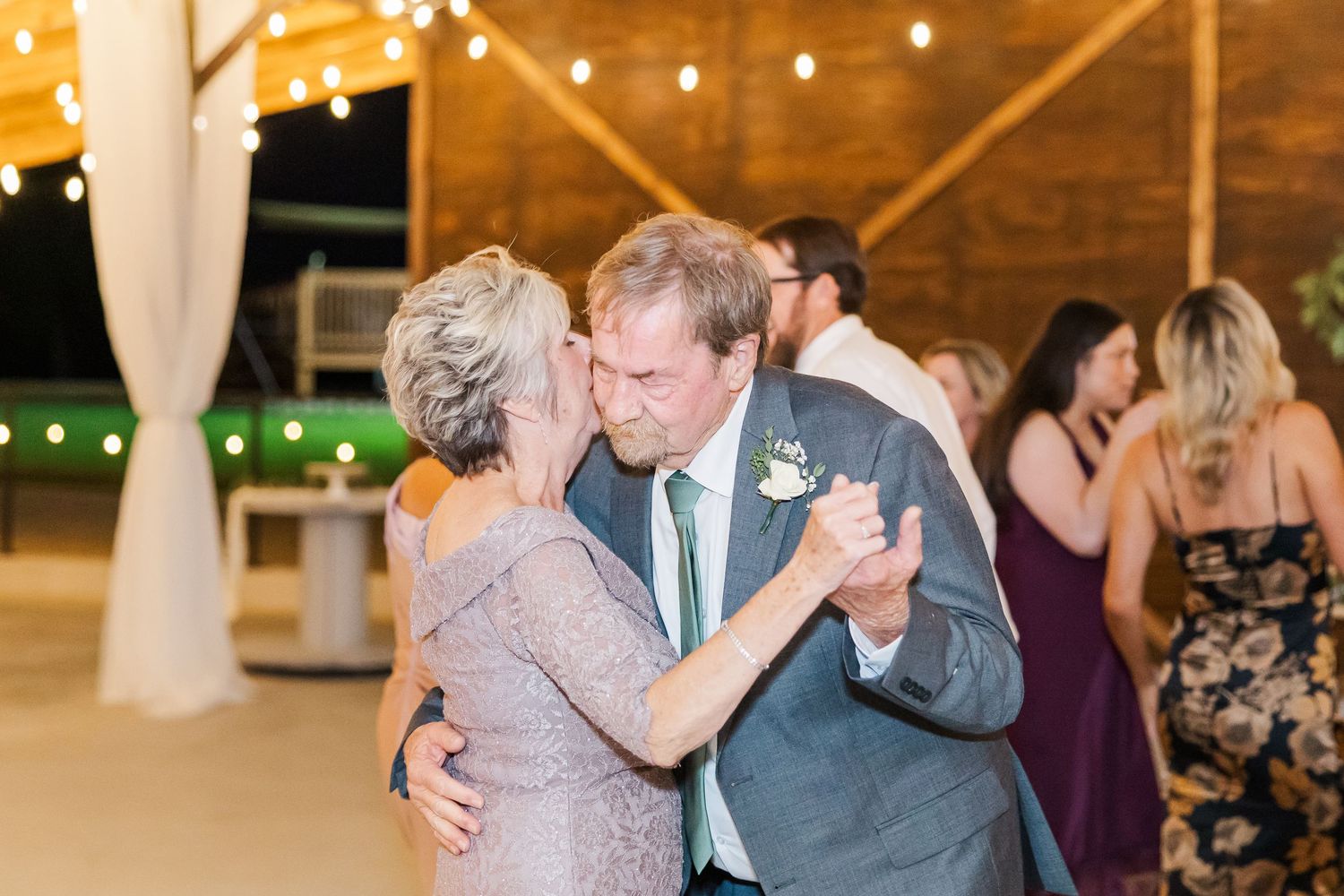 Elderly couple shares a romantic slow dance at a wedding reception under warm string lights.