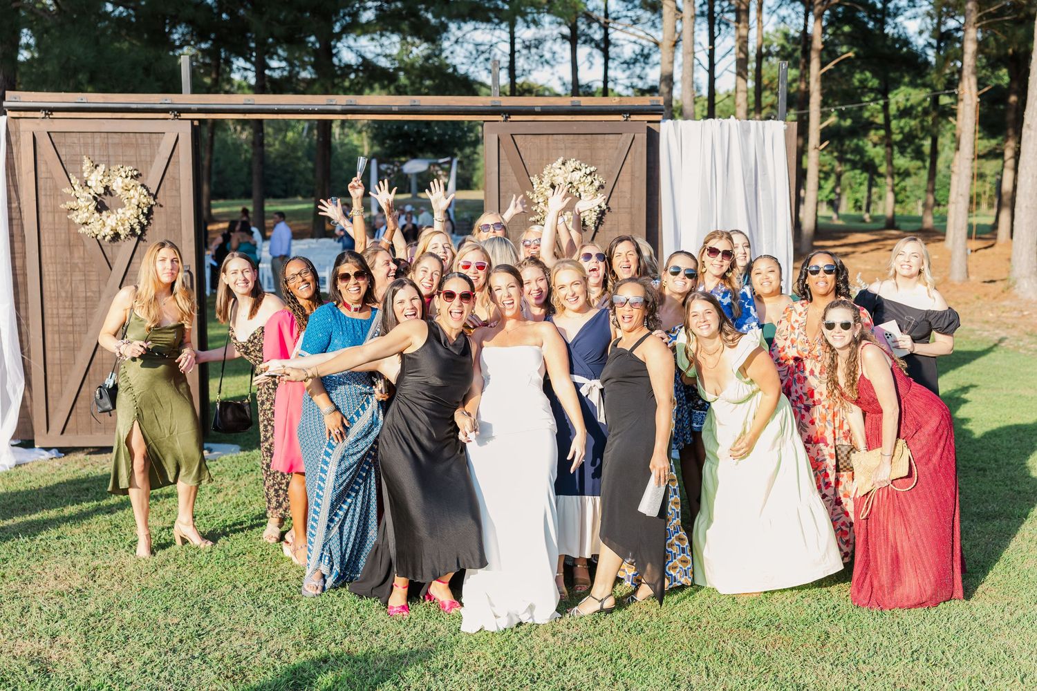 Bride poses with a large group of smiling female wedding guests outdoors near barn doors.