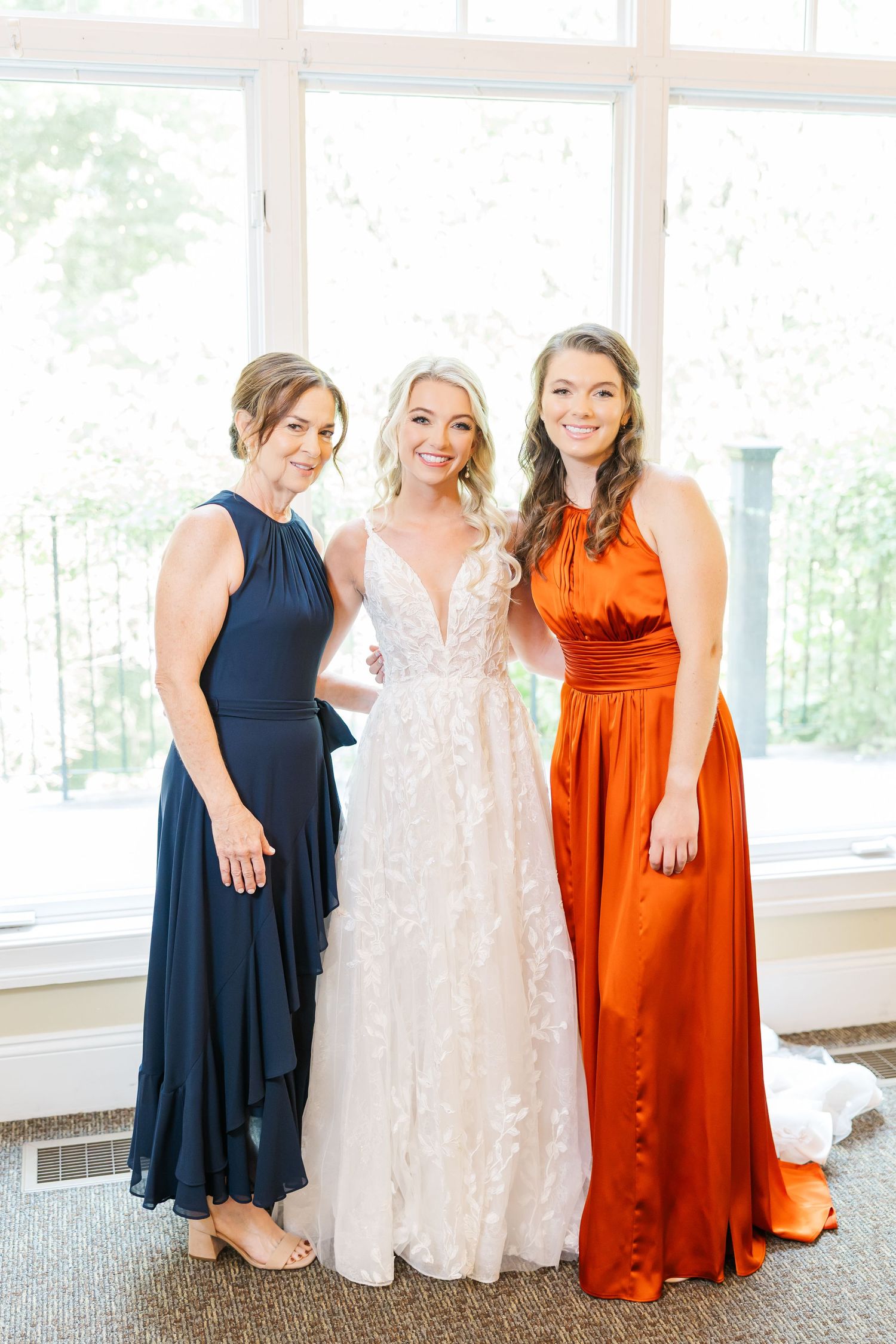 Bridesmaids in navy and orange dresses pose with bride in lace gown by bright windows.