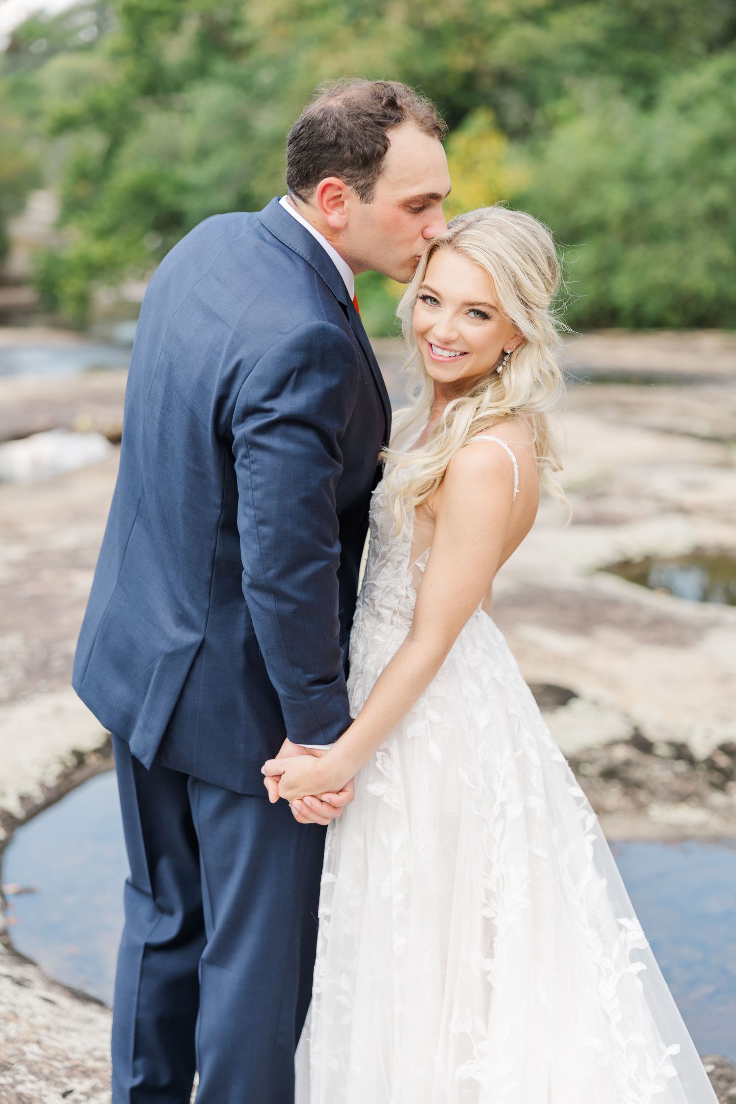 Wedding couple shares intimate moments on a rocky riverbank in elegant attire.