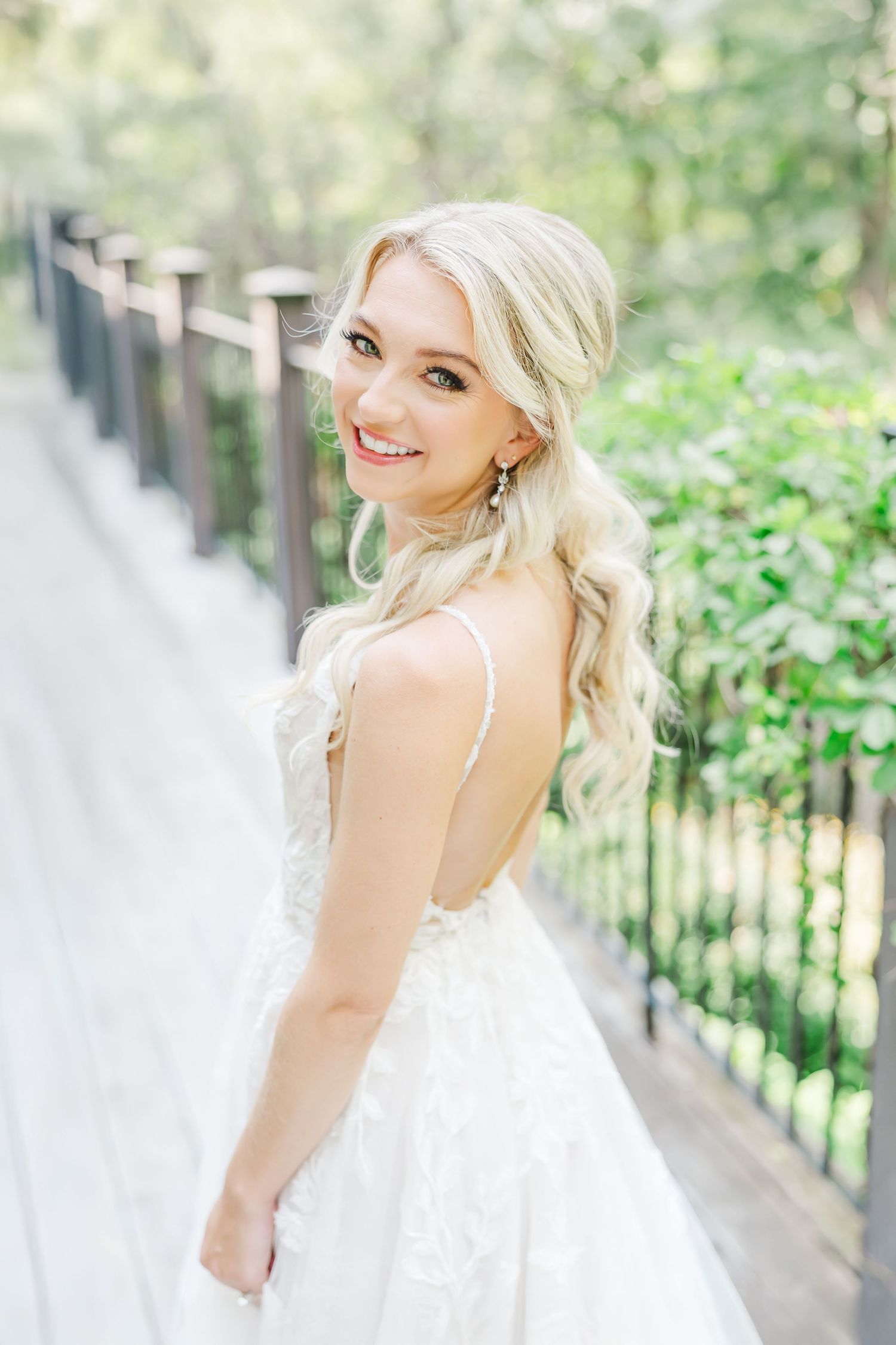 An elegant bride in a backless wedding gown poses on a pathway with an iron railing.