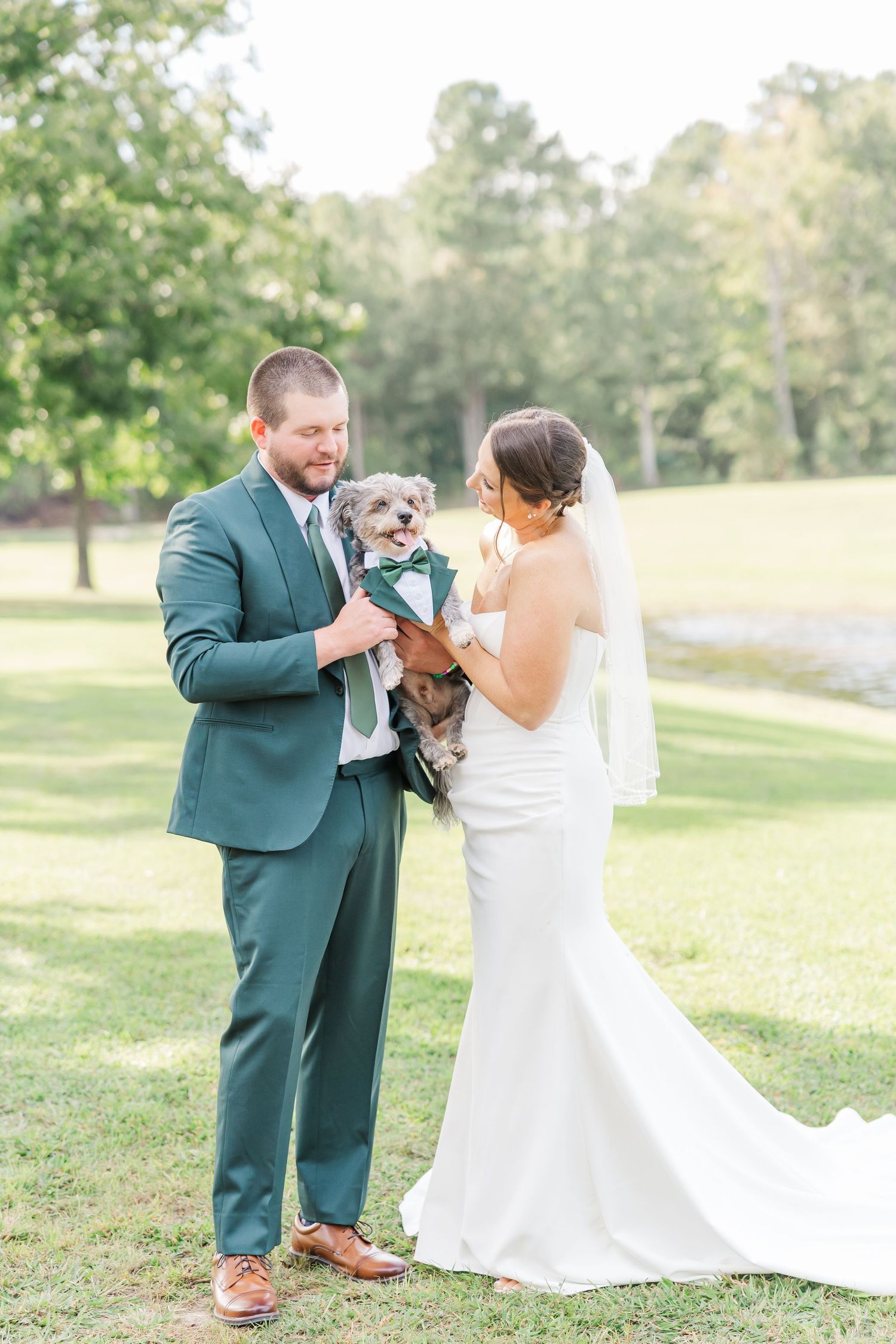 A couple in wedding attire shares a joyful moment with their dog in a sunny outdoor setting.