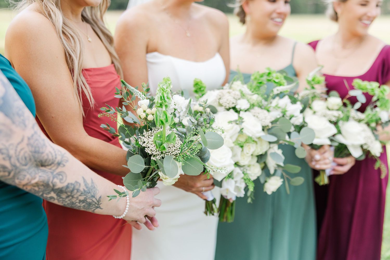 Wedding party members in colorful dresses hold elegant white and green floral bouquets during outdoor ceremony.