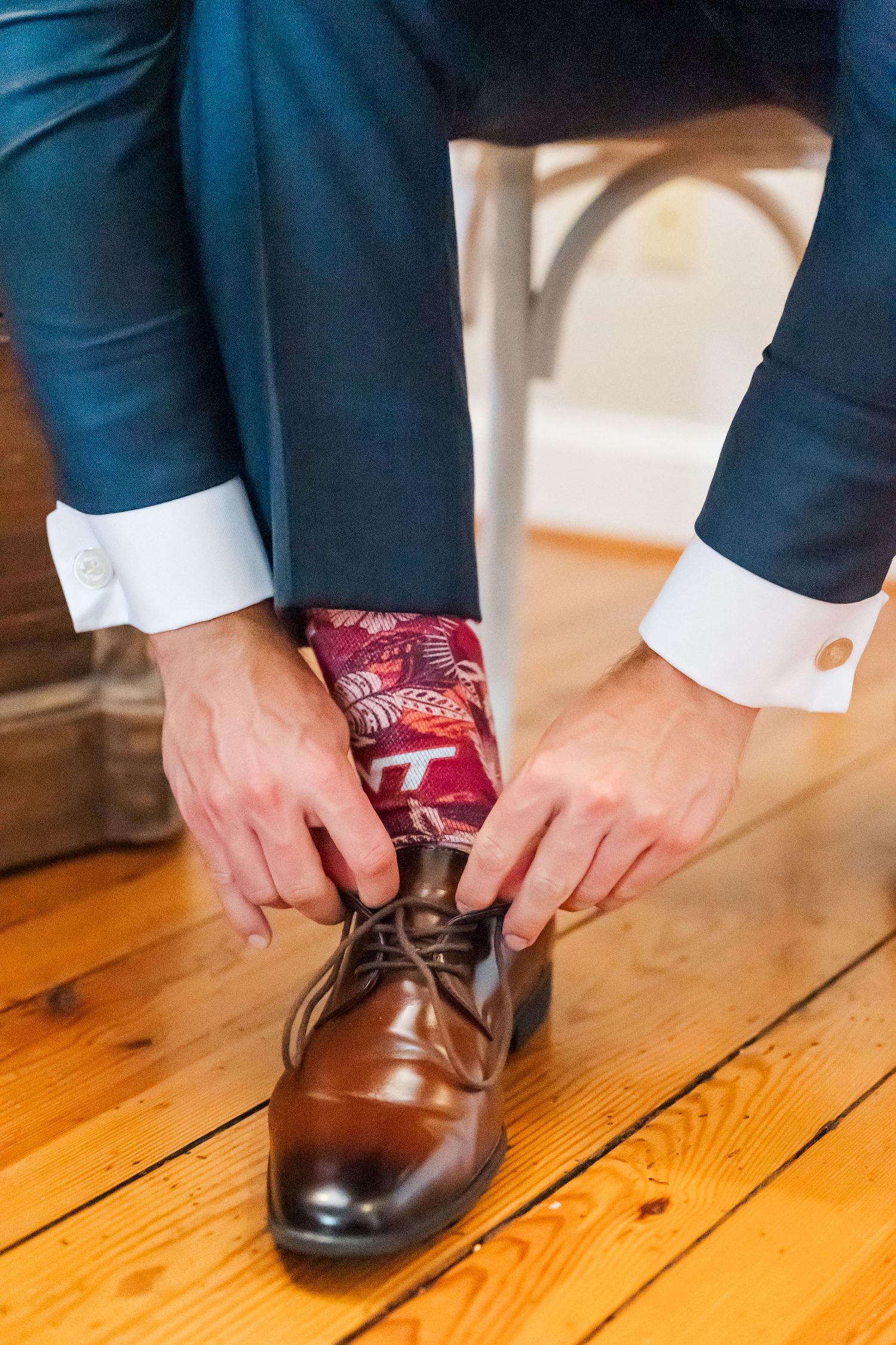 Close-up of groom wearing brown leather dress shoes and burgundy patterned socks with navy suit on wooden floor.