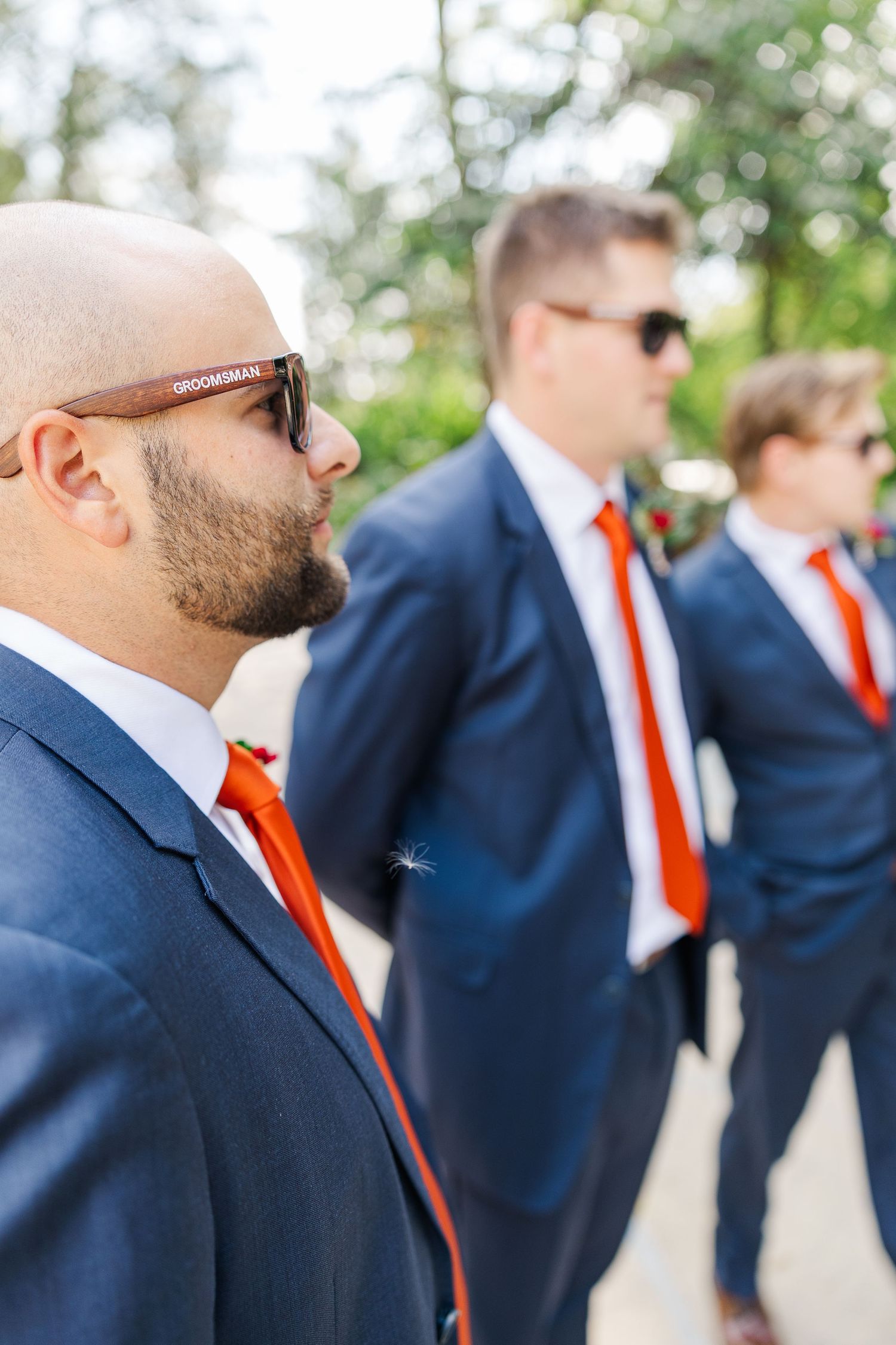 Groomsmen in matching navy suits and orange ties stand in a diagonal line at an outdoor wedding.