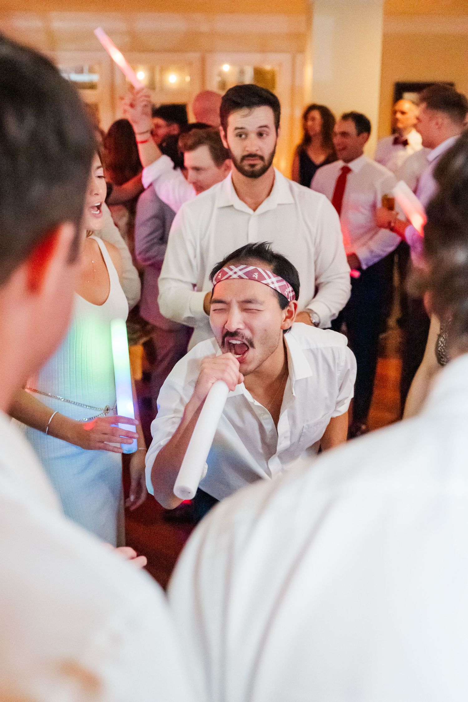 Wedding reception dance floor filled with energetic guests celebrating in white attire under colorful party lights.