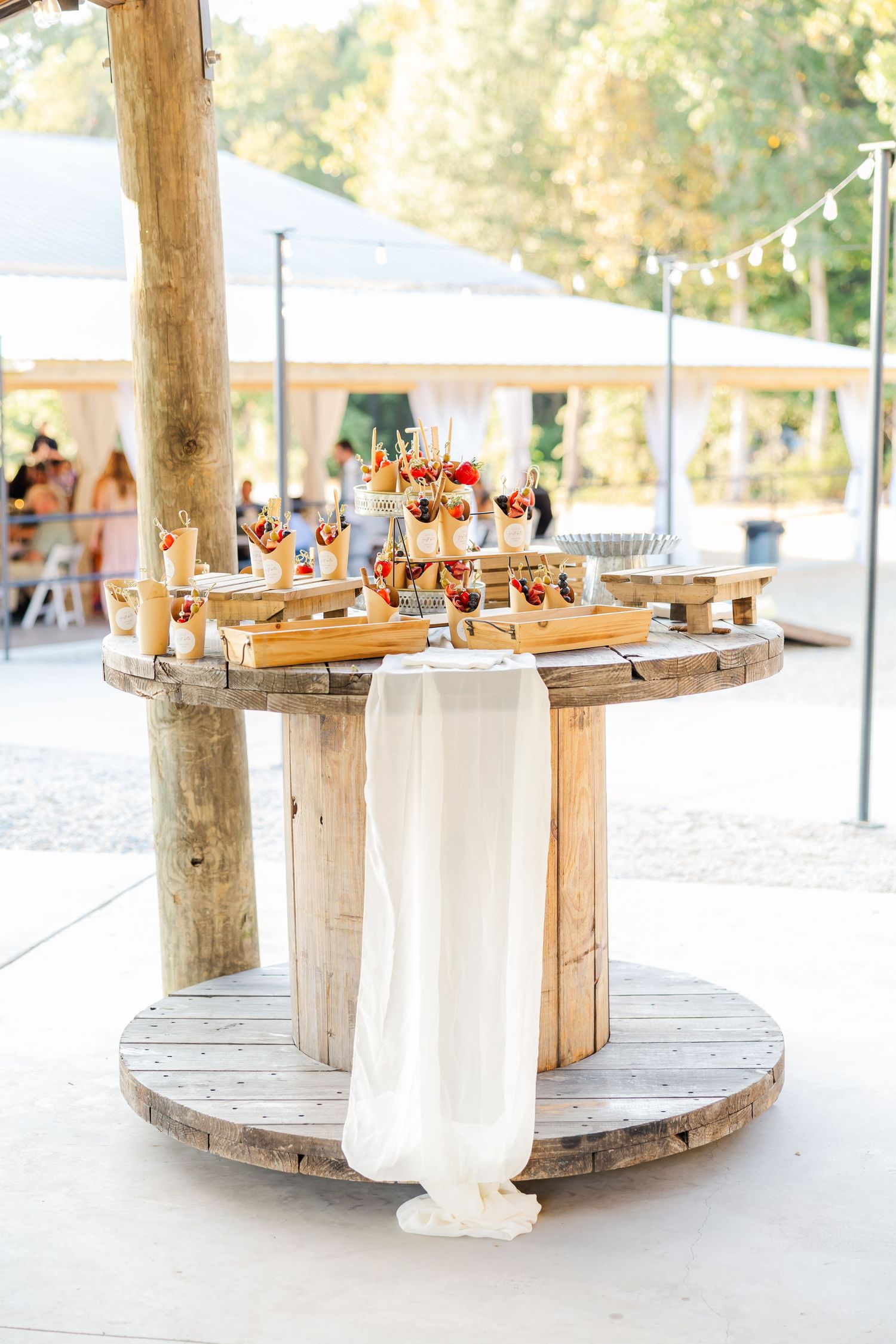 Rustic wooden spool table decorated with elegant desserts and white fabric draping at an outdoor wedding reception.