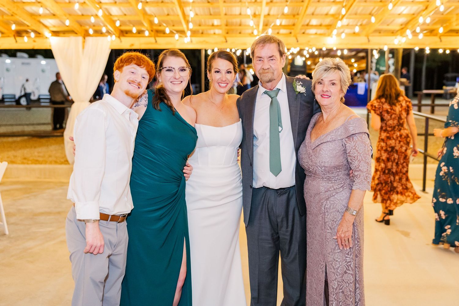 Family group photo at elegant wedding reception with string lights, bride in white gown with guests in formal attire.