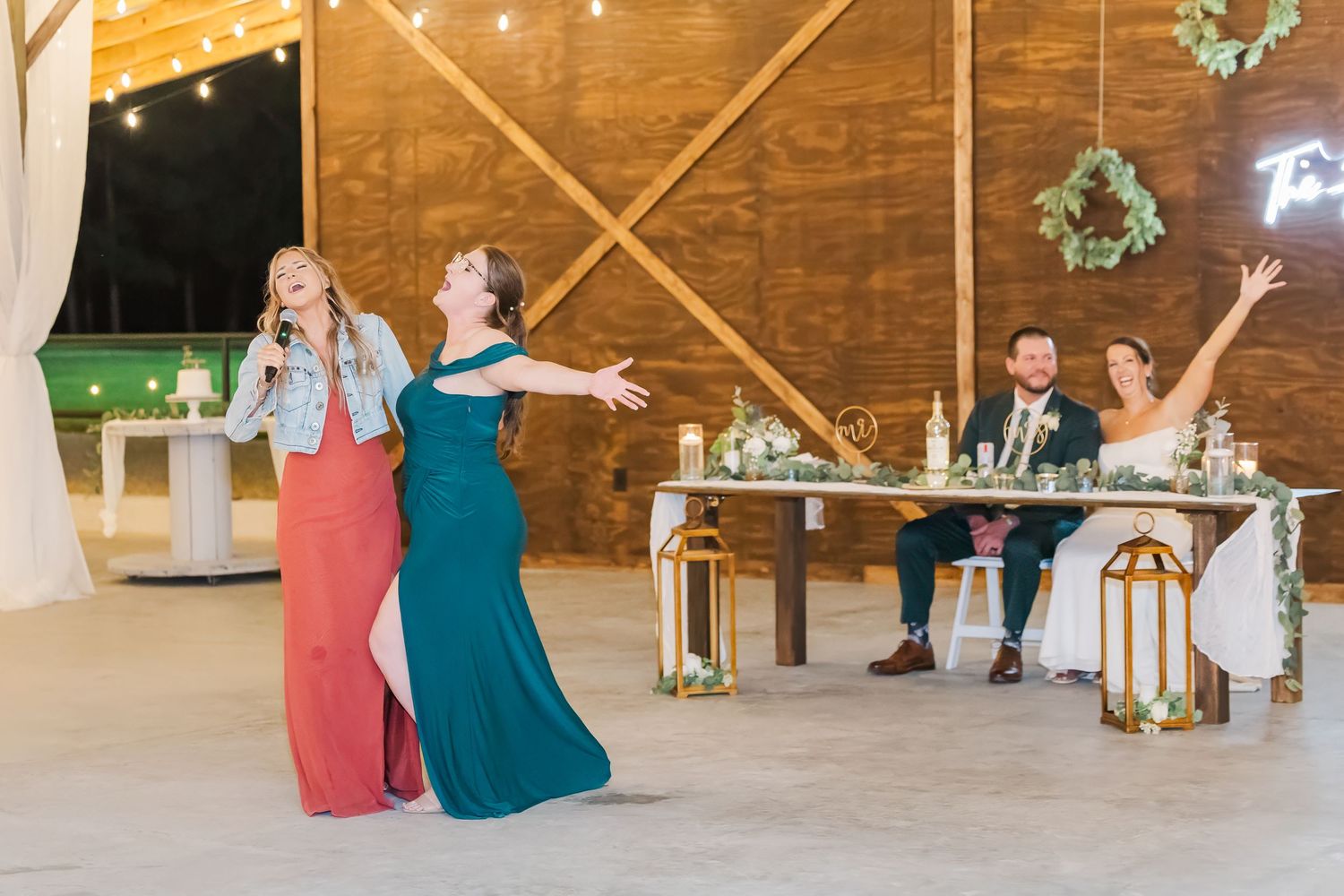 Two bridesmaids perform a fun speech while the newlywed couple celebrates joyfully at their rustic barn reception.