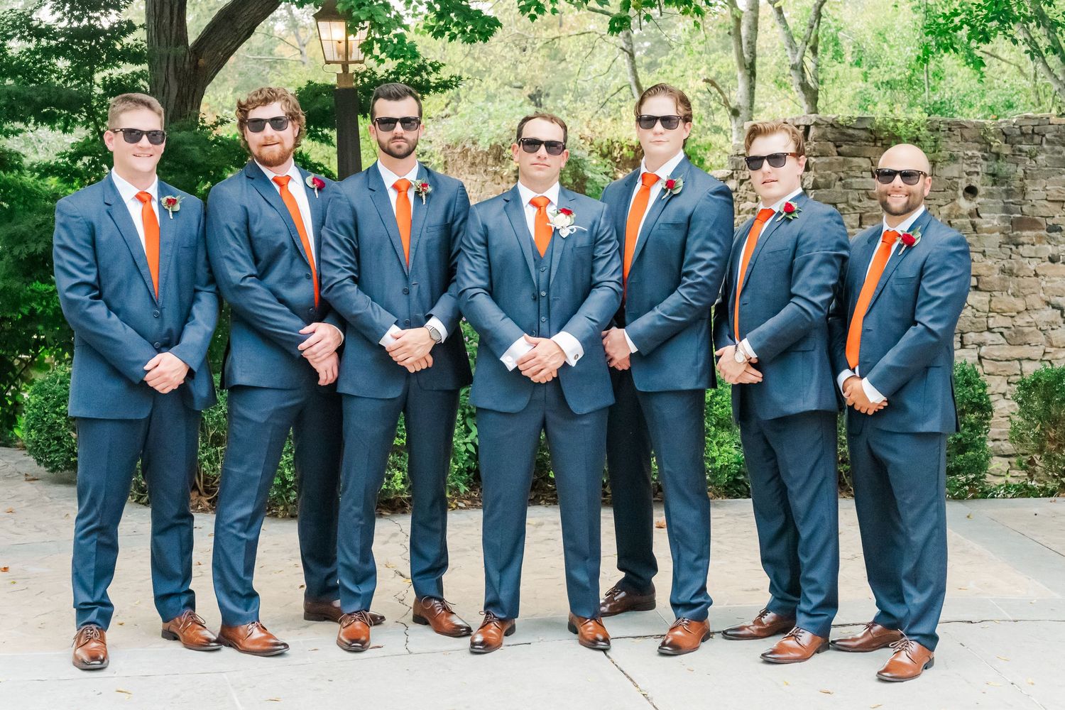 Groomsmen in navy suits and orange ties show off their brown leather shoes while seated on stone steps.