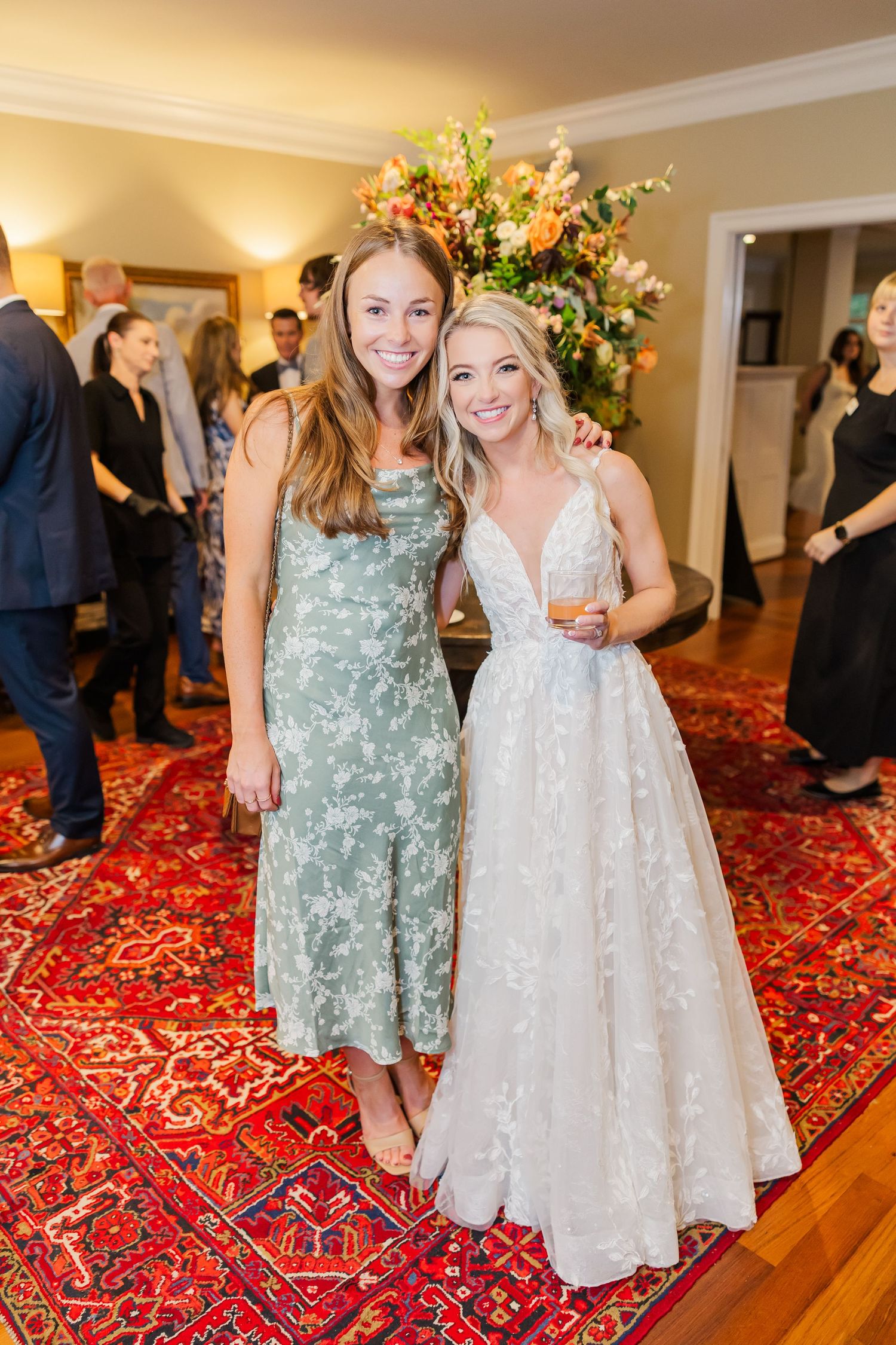 Wedding party members in sage green and white dresses pose on a patterned red carpet.
