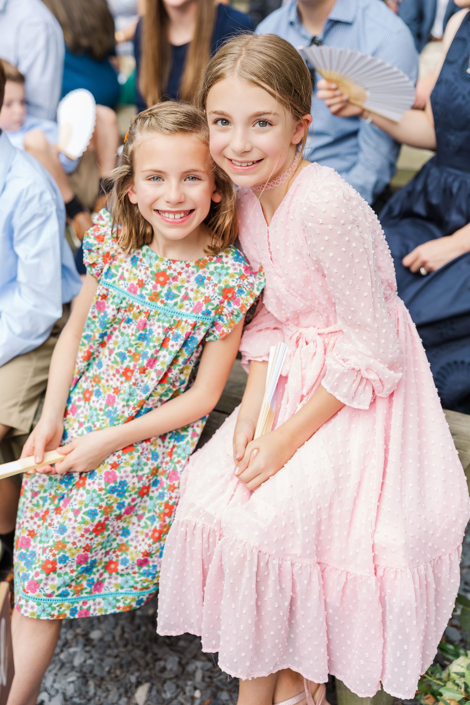 Two children wearing floral and pink dresses sit together at an outdoor event.