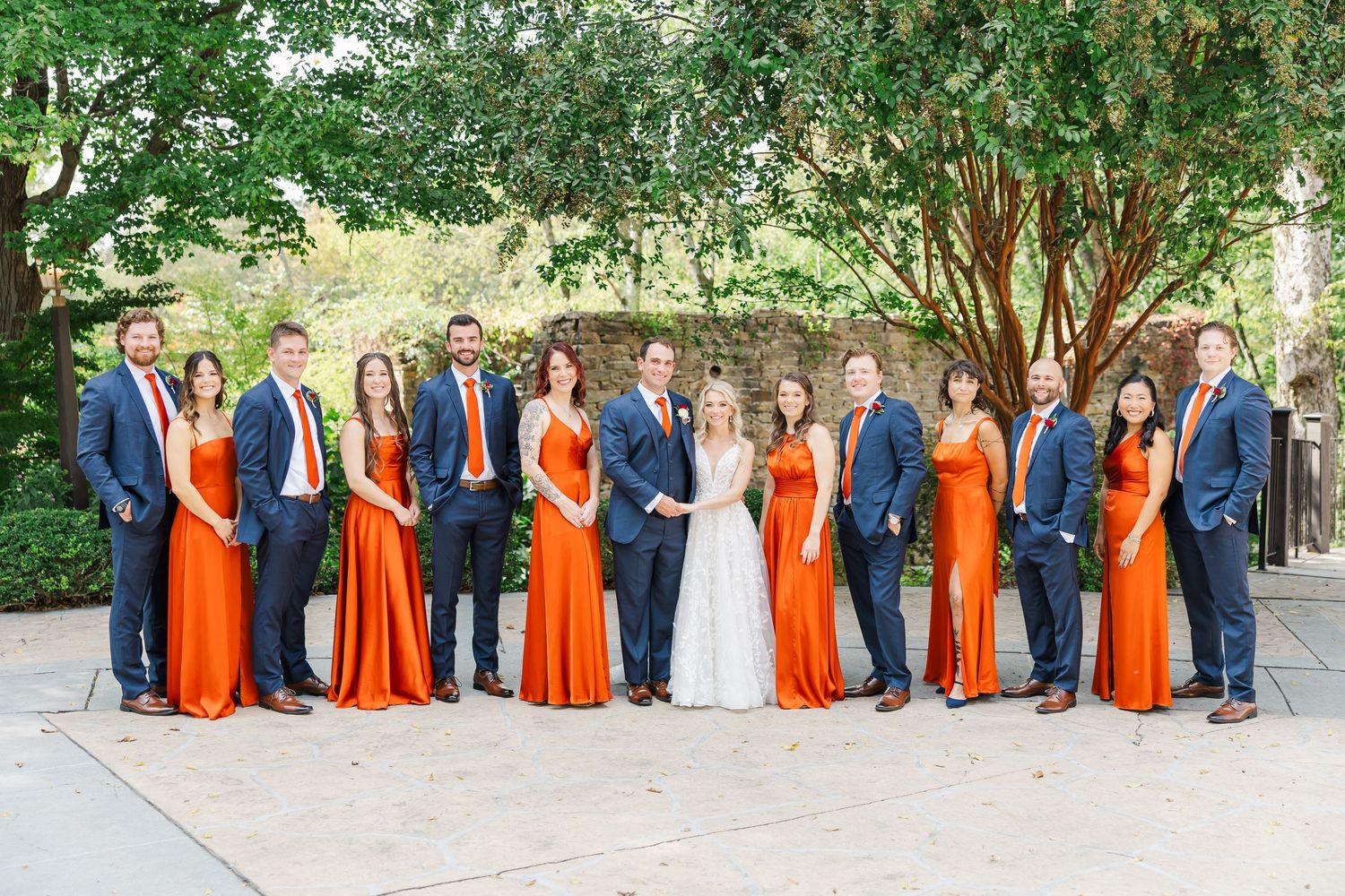 Wedding party in navy blue and bright orange attire poses together in an outdoor garden setting.