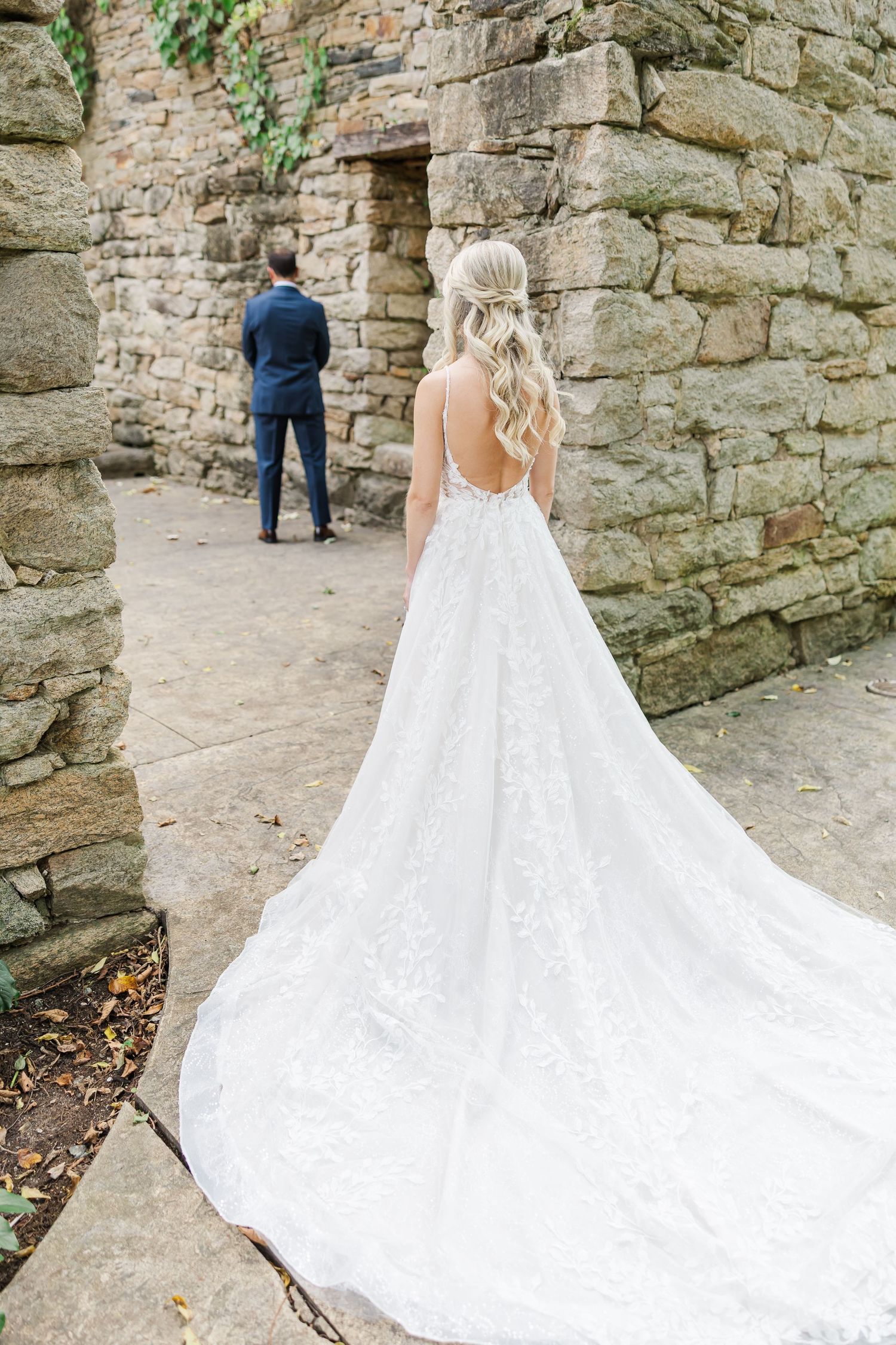 A wedding couple stands near stone walls while the bride's elegant white lace gown with long train flows behind her.