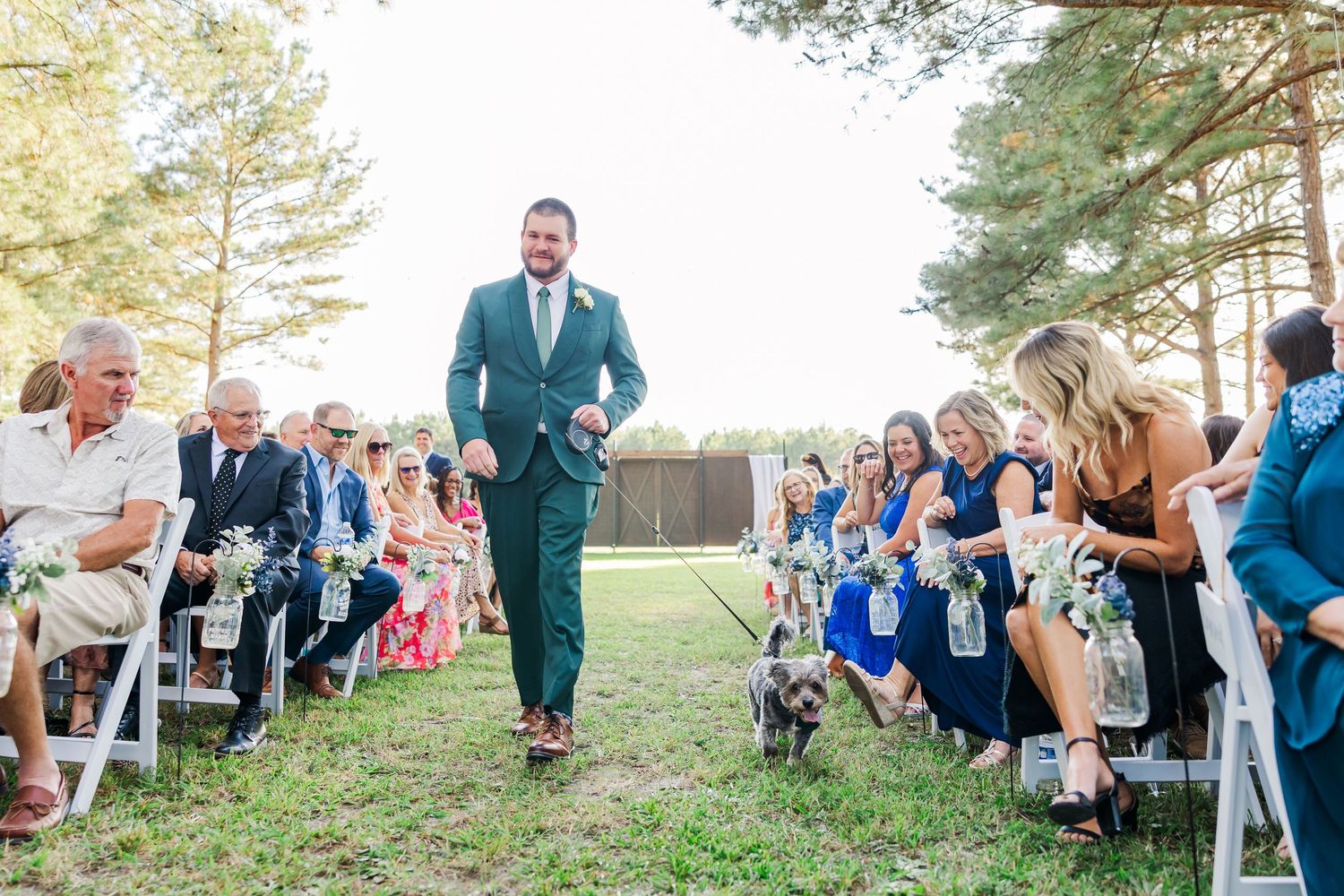 Groom in green suit walks down outdoor wedding aisle with small dog, guests seated in white chairs among tall pine trees.