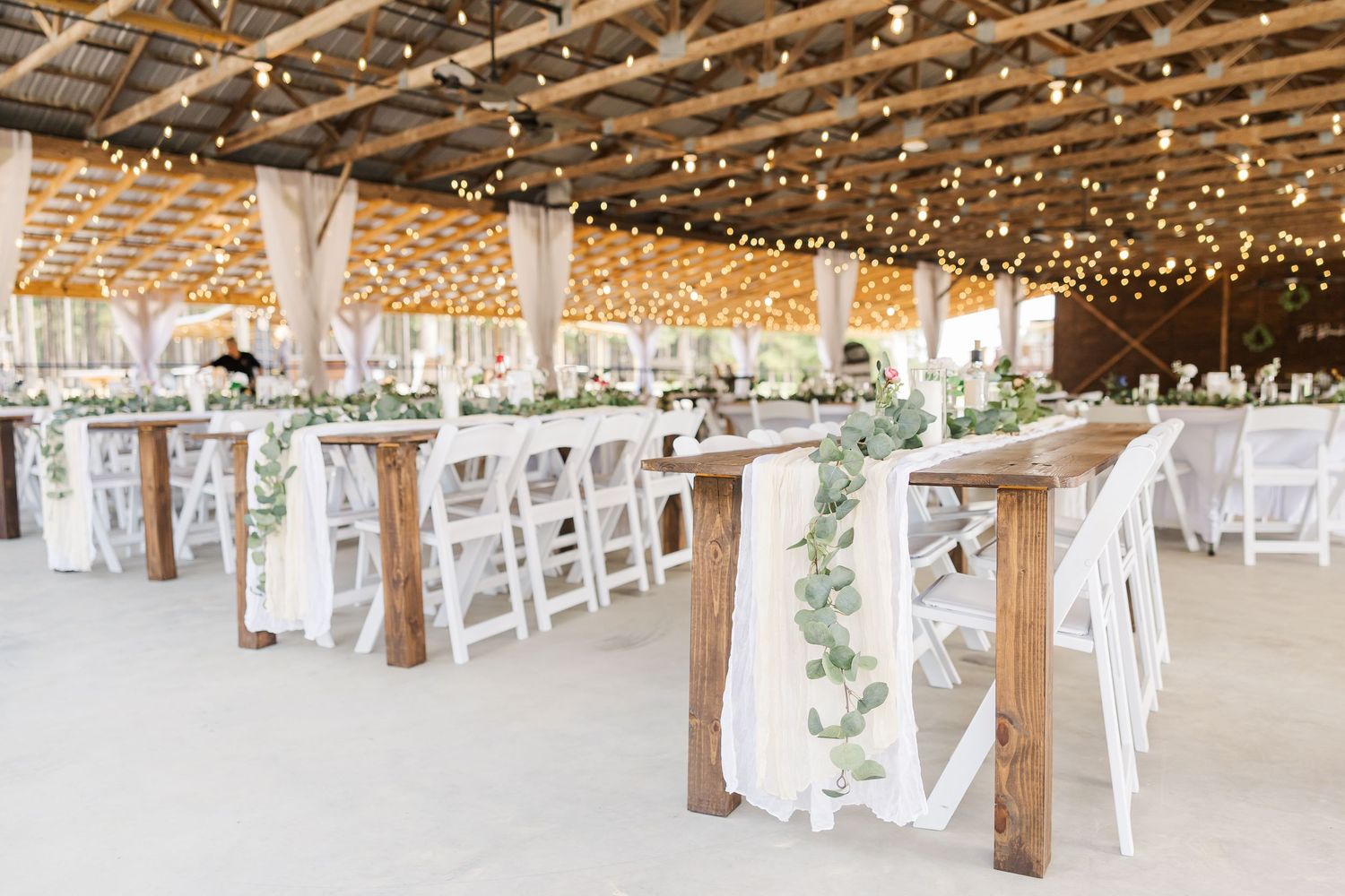 Rustic barn wedding reception with string lights, wooden tables, white chairs, and eucalyptus garland centerpieces.