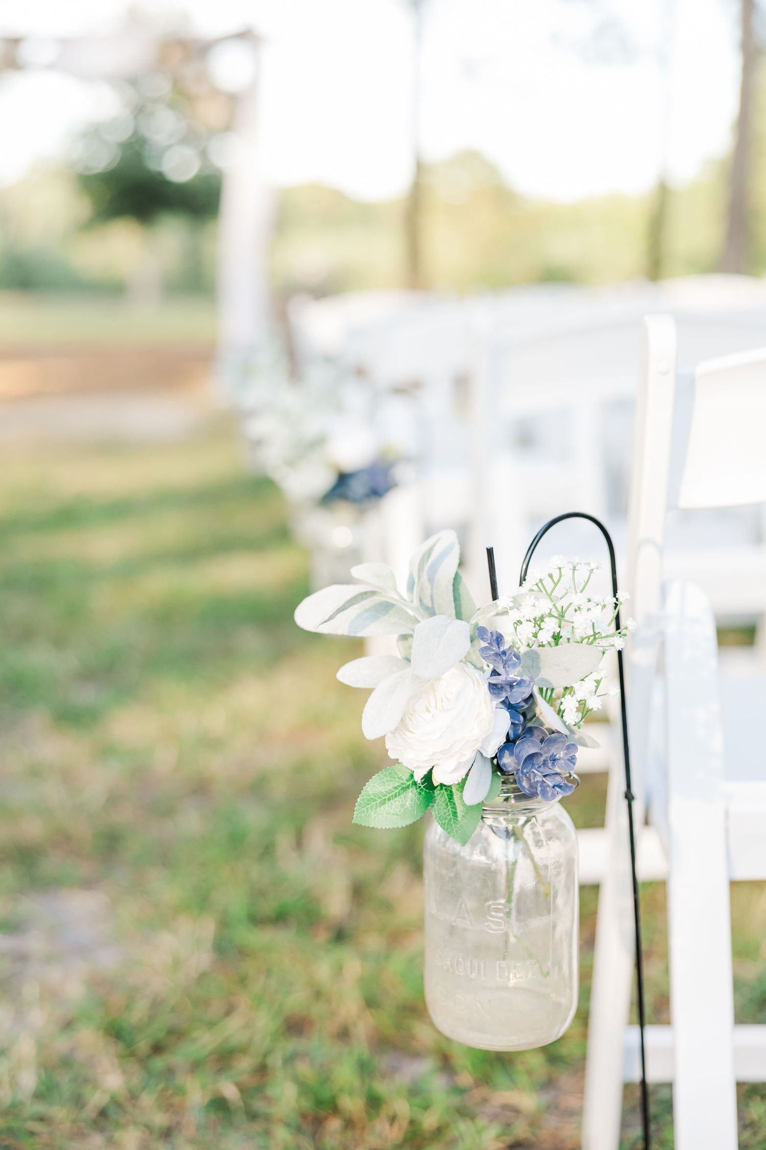 Mason jar with white and blue flowers hanging on shepherd hook along wedding ceremony aisle with white chairs.