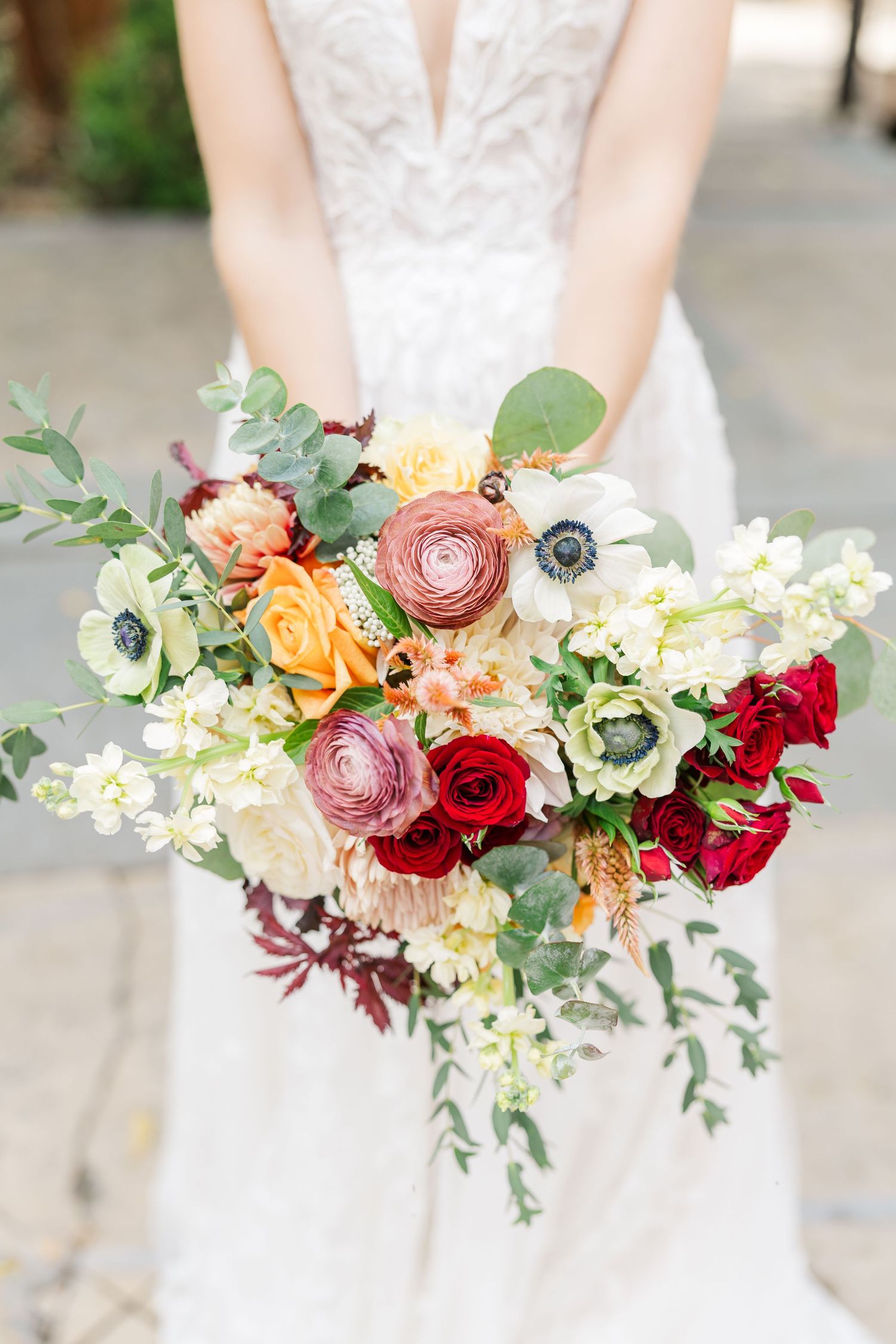 A stunning bridal bouquet featuring pink ranunculus, red roses, white orchids, and eucalyptus greenery against a white dress.