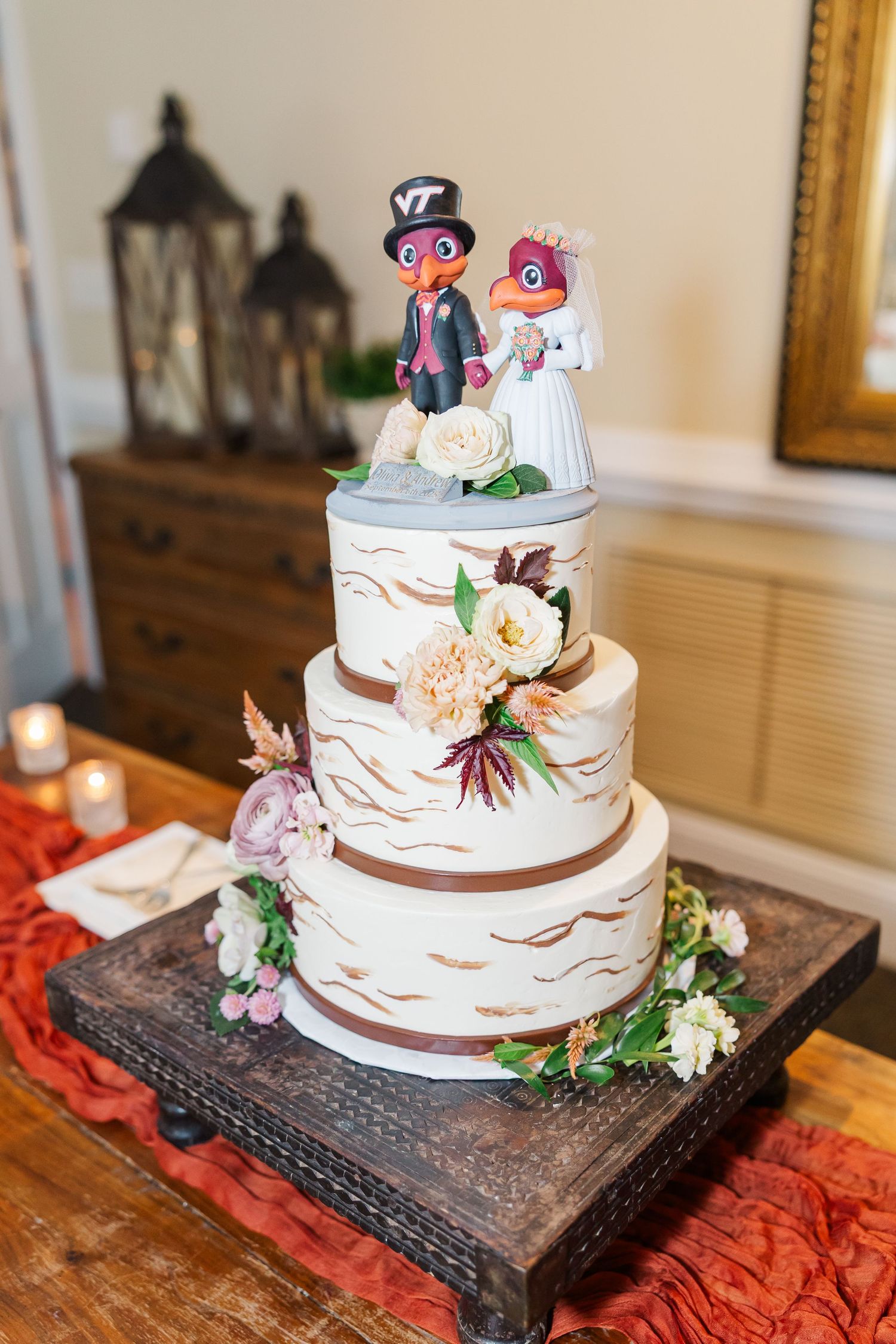 Three-tiered white wedding cake decorated with fresh flowers and cute animal cake toppers sits on dark wooden stand.