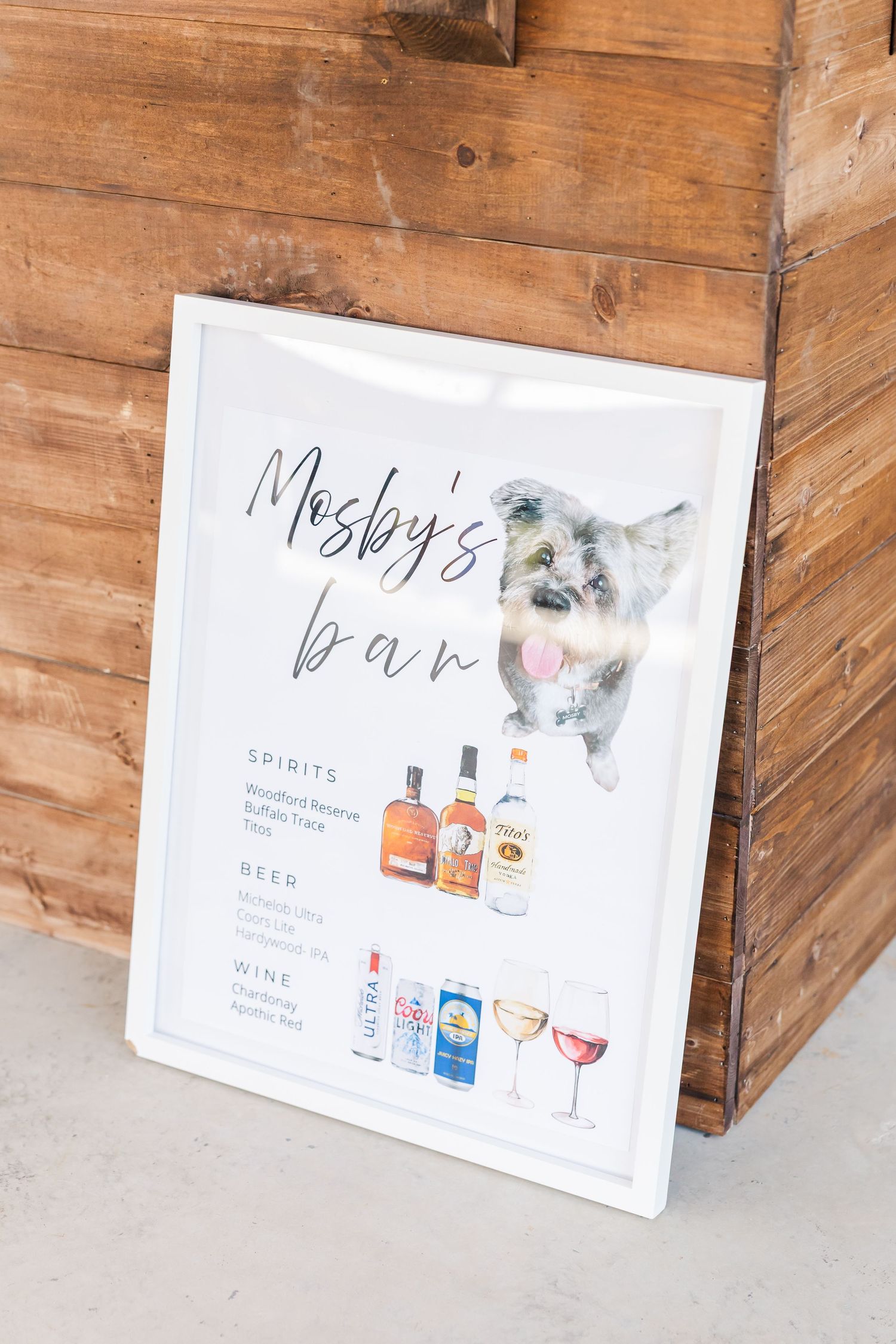 Mosby's Bar sign with watercolor dog illustration listing spirits, beer, and wine options, leaning against rustic wood wall.
