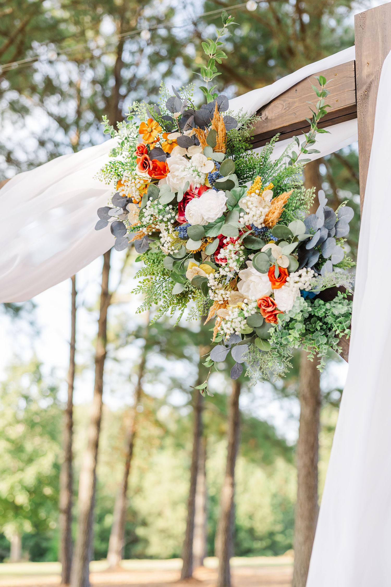 Colorful wedding arch floral arrangement with orange, red, and white flowers, eucalyptus, and white draped fabric outdoors.