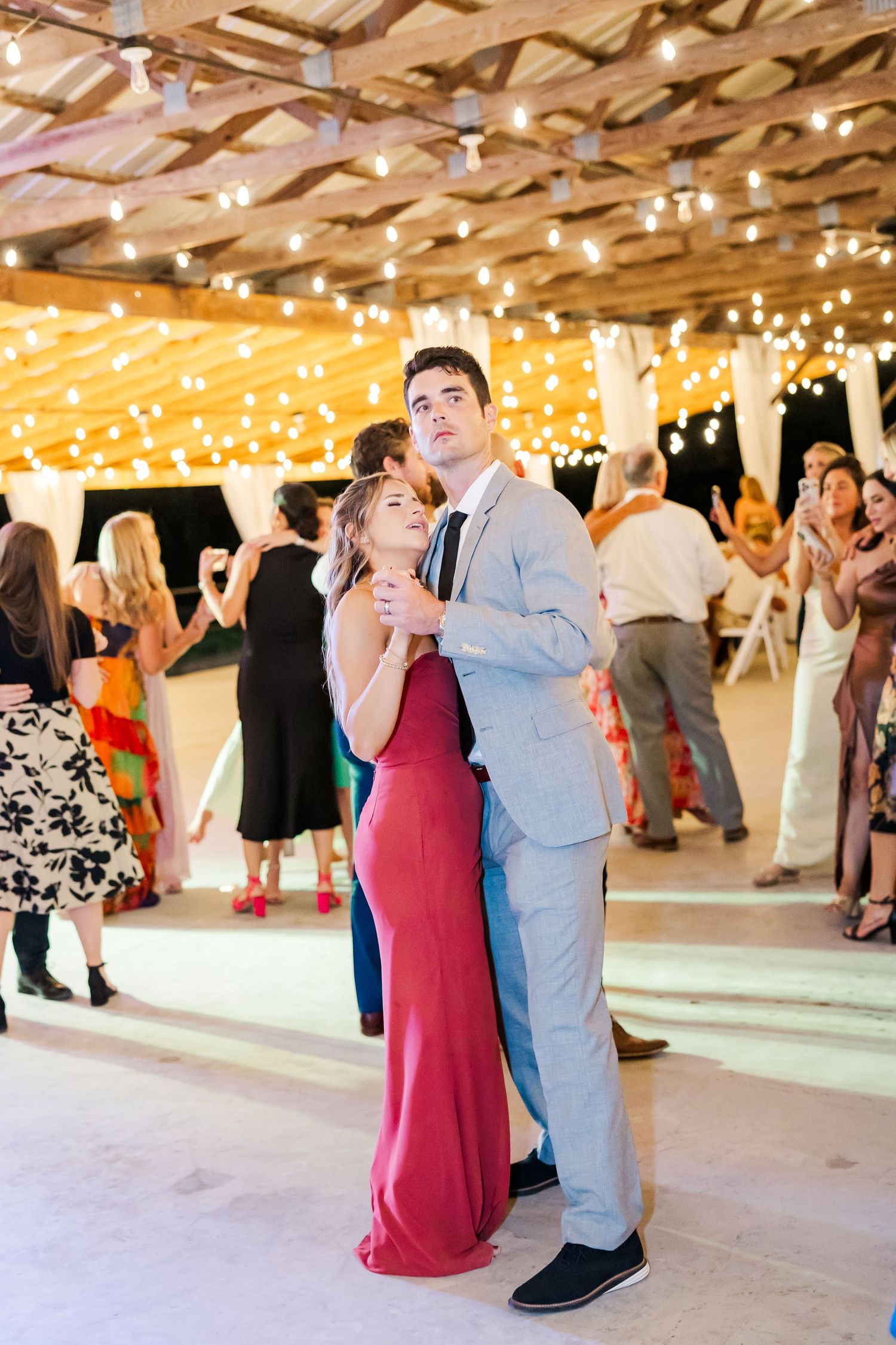 A couple shares a romantic dance in a warmly lit wedding reception venue decorated with string lights overhead.