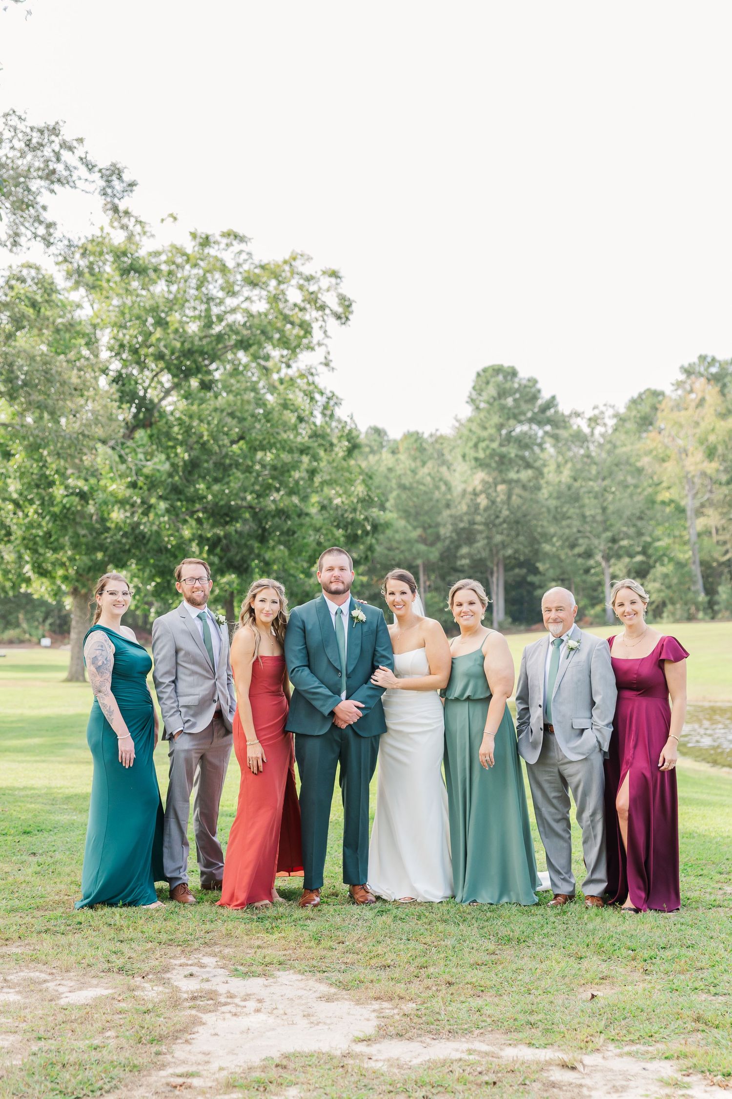 Wedding party in colorful dresses and suits pose together outdoors with lake and trees in background.