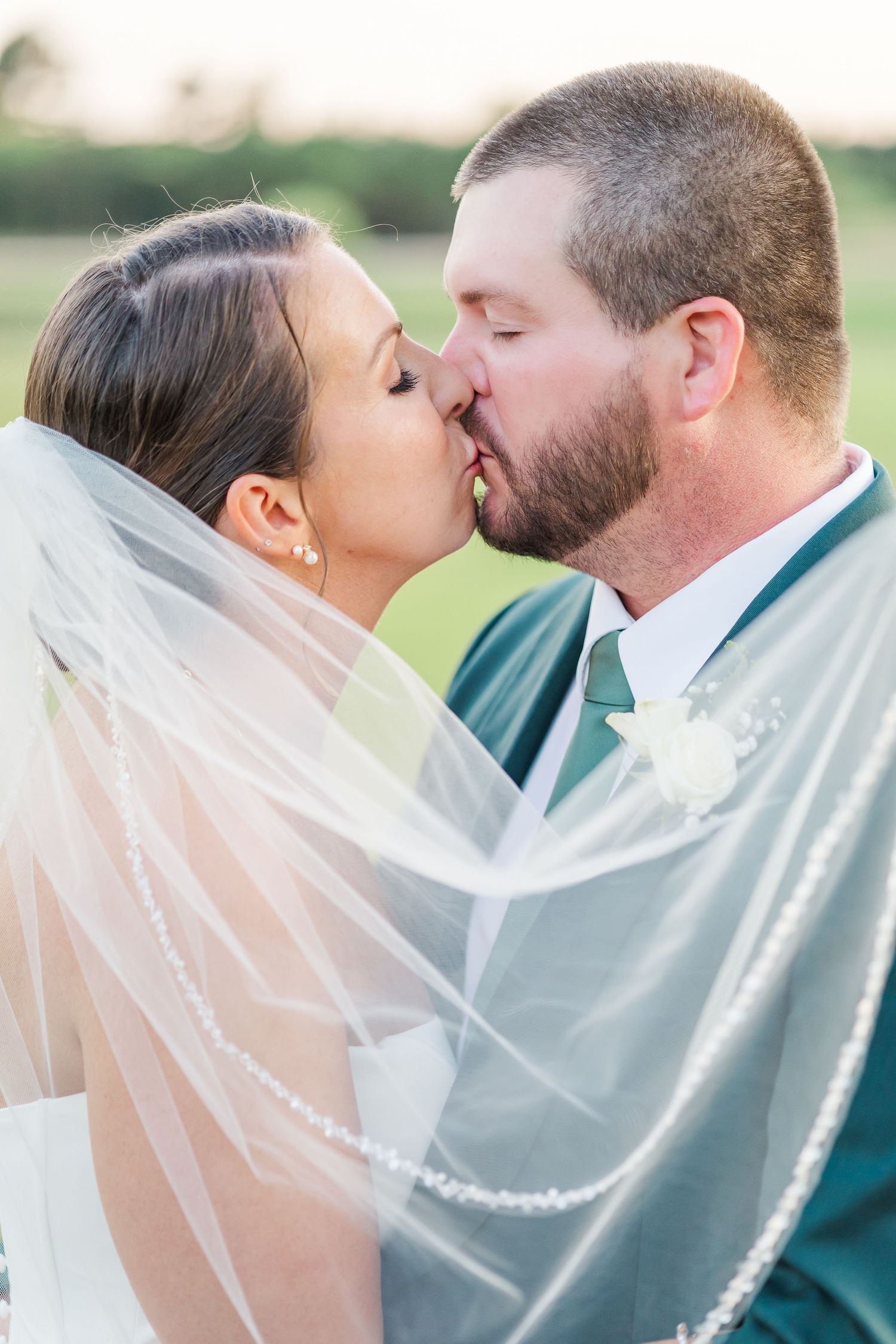 A romantic wedding kiss captured outdoors with the bride wearing a flowing veil and groom in a teal colored suit.