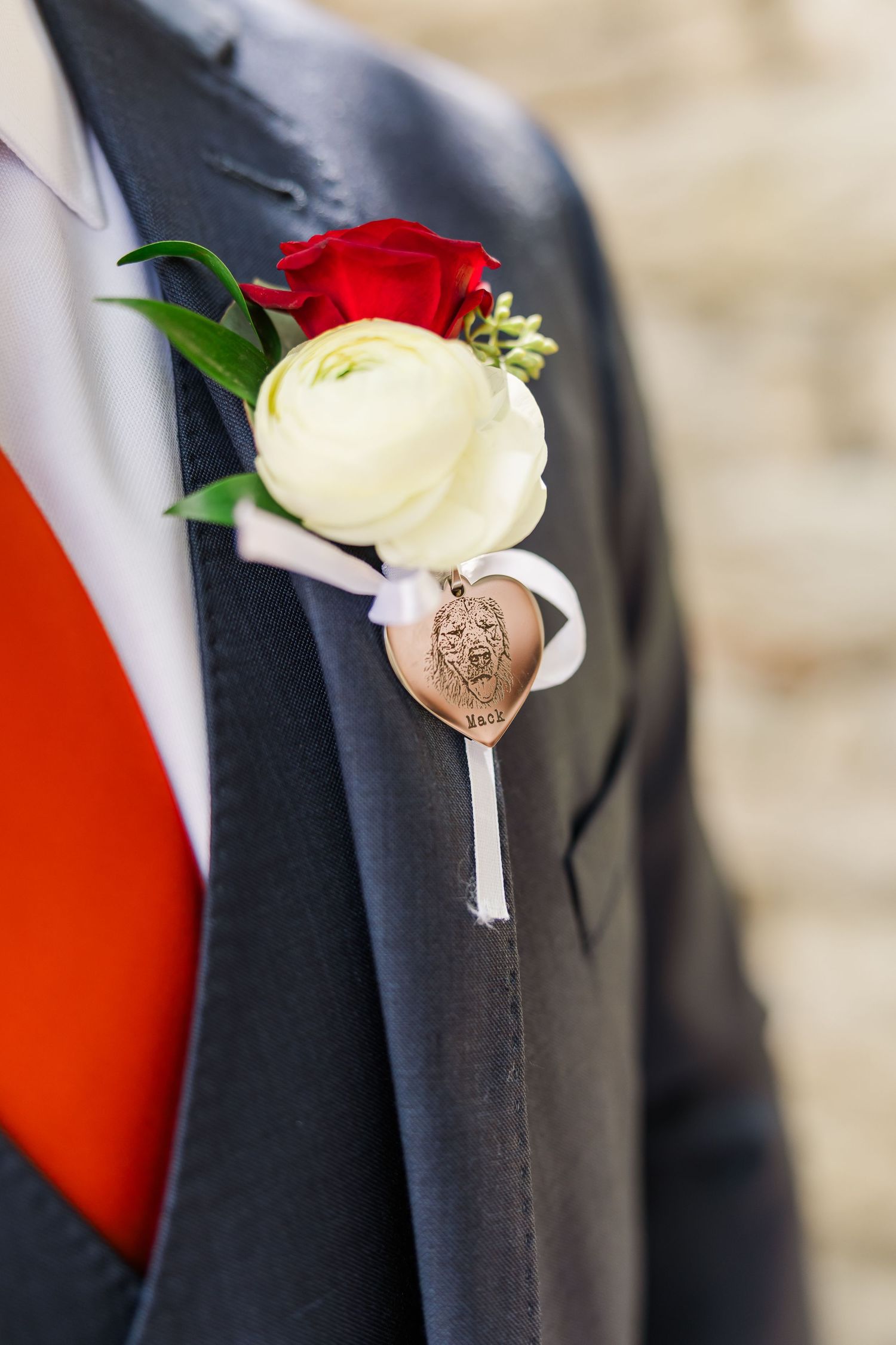 A wedding boutonniere with red and white roses adorned with a heart-shaped charm pinned to a dark suit jacket.