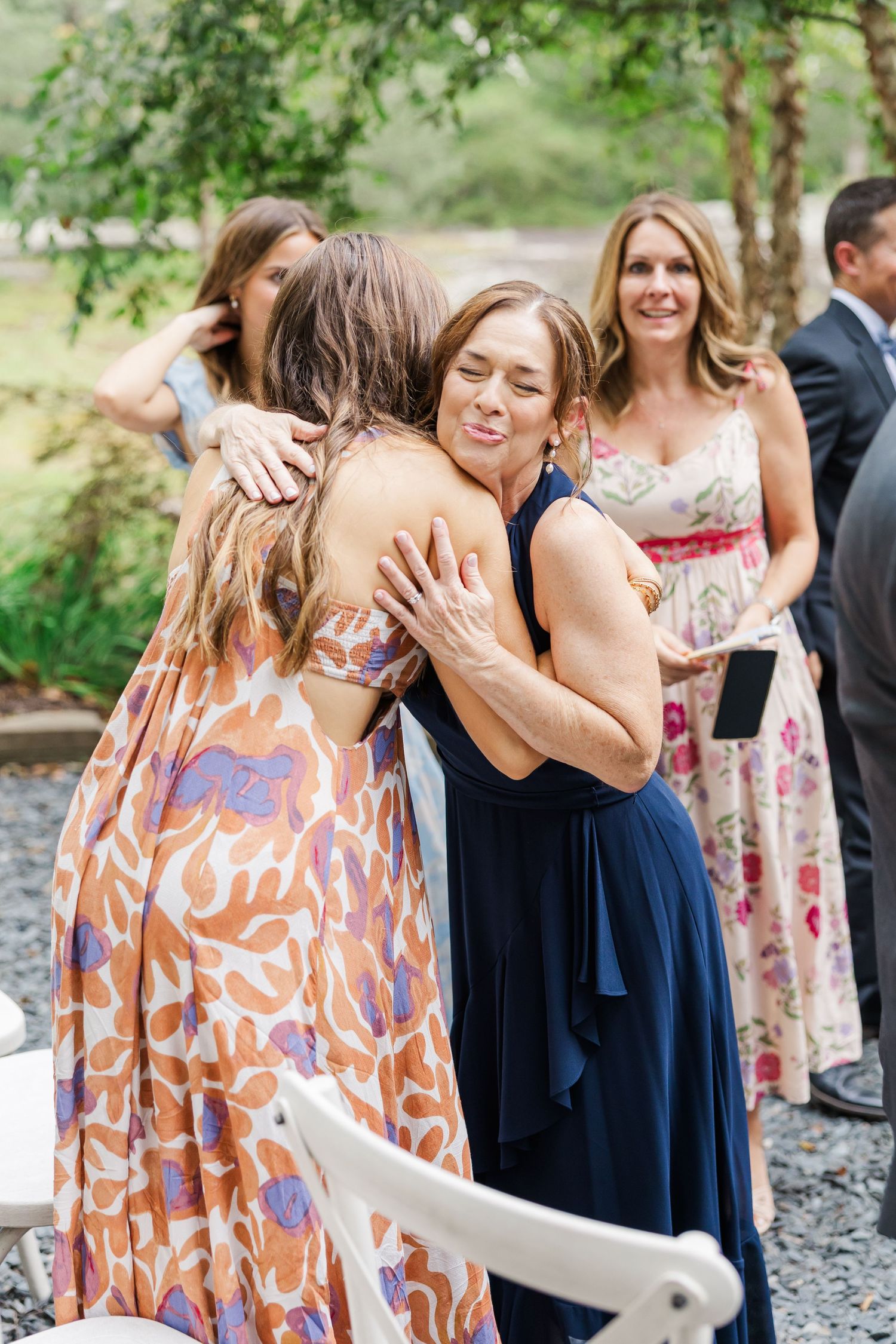 Wedding guests in navy and floral dresses share emotional hugs and embraces during an outdoor celebration.