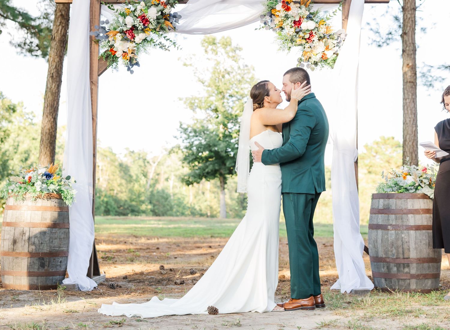 Bride and groom share first kiss under floral arch with white draping and barrel accents at outdoor wedding ceremony.