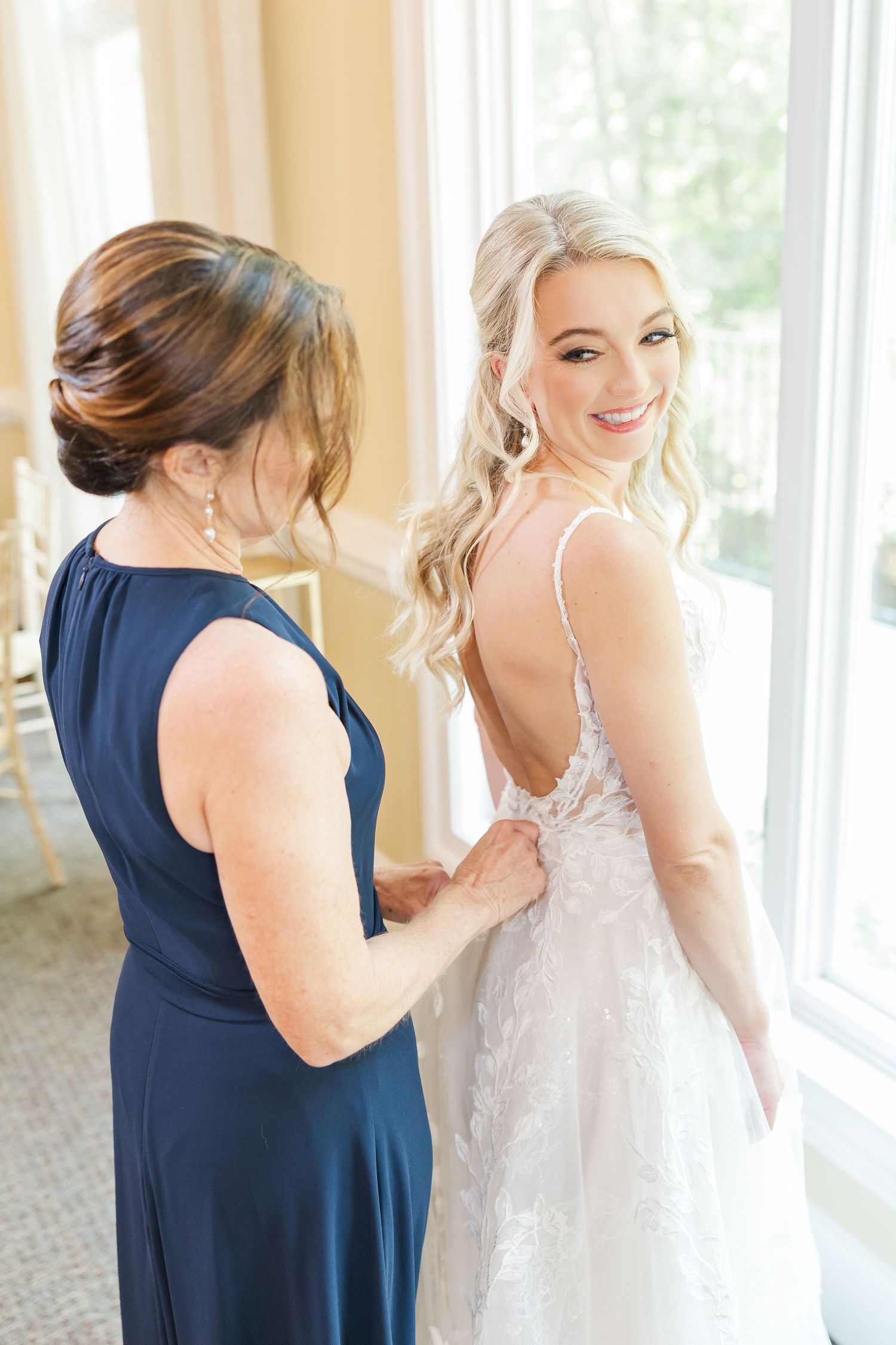 Wedding party helps bride with final dress preparations in bright sunlit room.