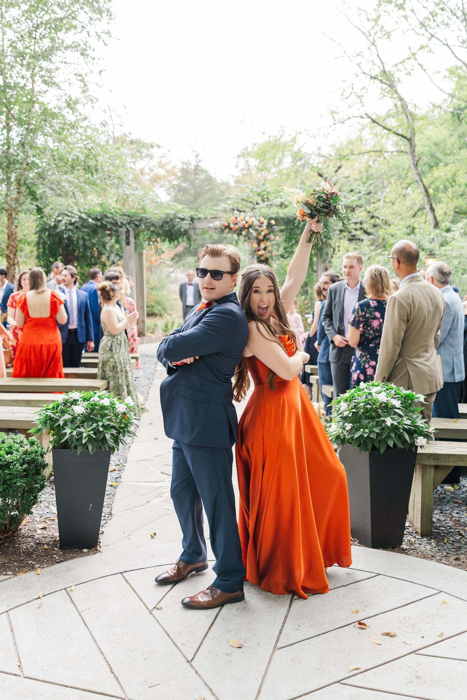 Wedding guests dance and celebrate on an outdoor patio with large decorative planters and greenery.