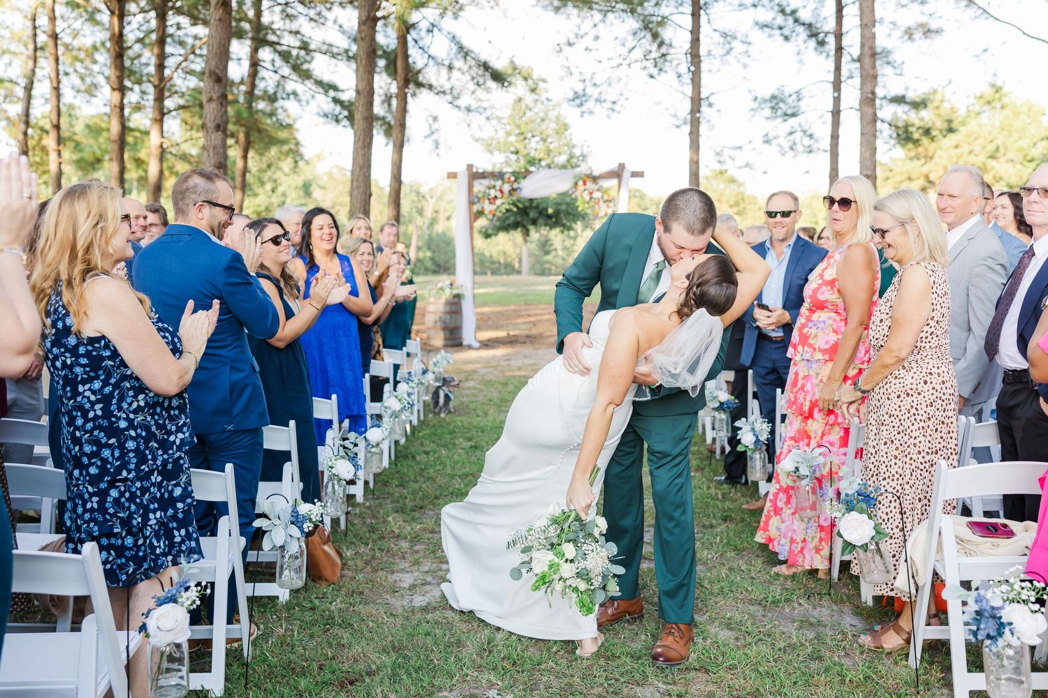 Bride and groom share a romantic kiss while walking down the aisle at an outdoor wedding ceremony with guests applauding.