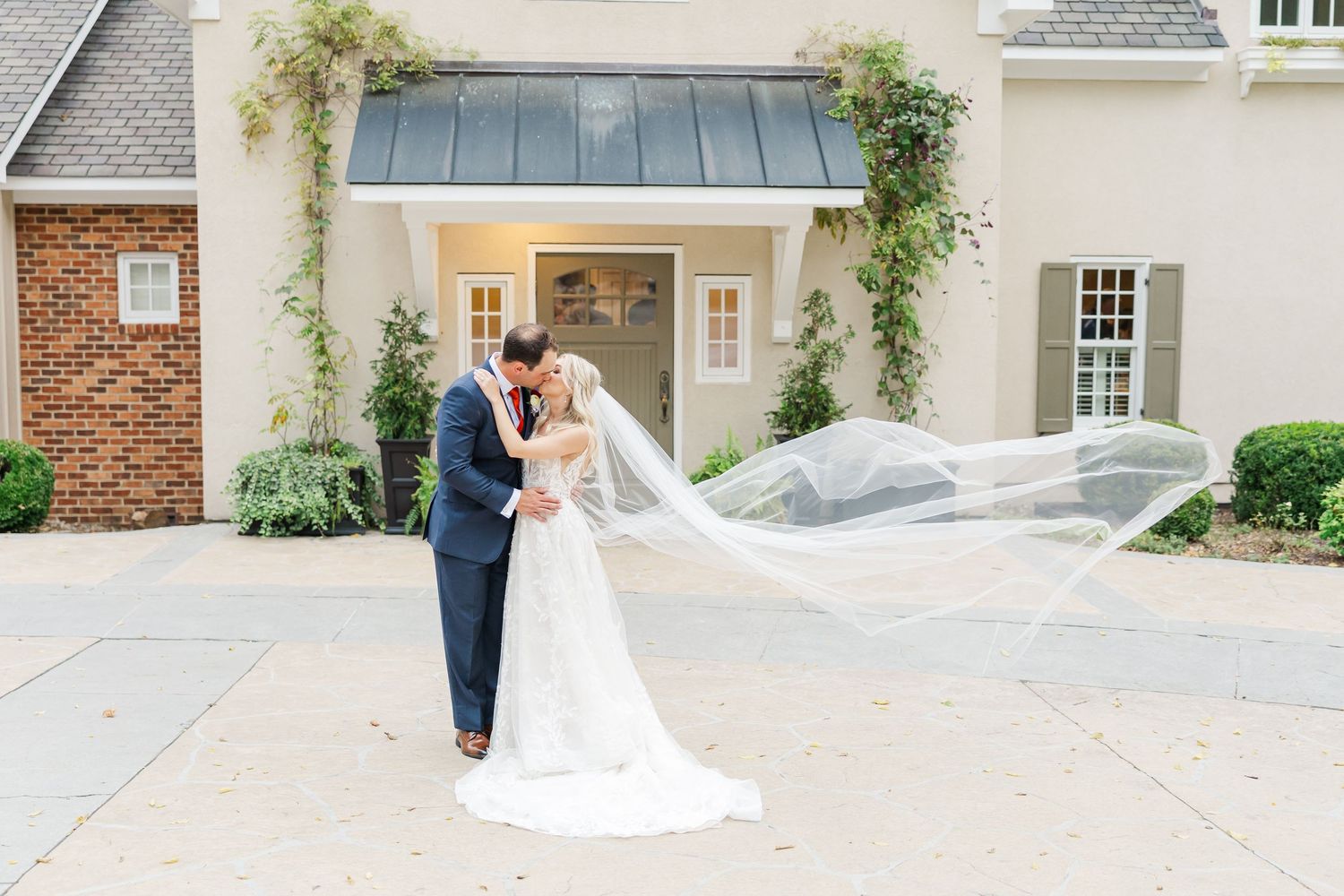 Elegant wedding couple poses outside stately brick mansion with climbing vines.