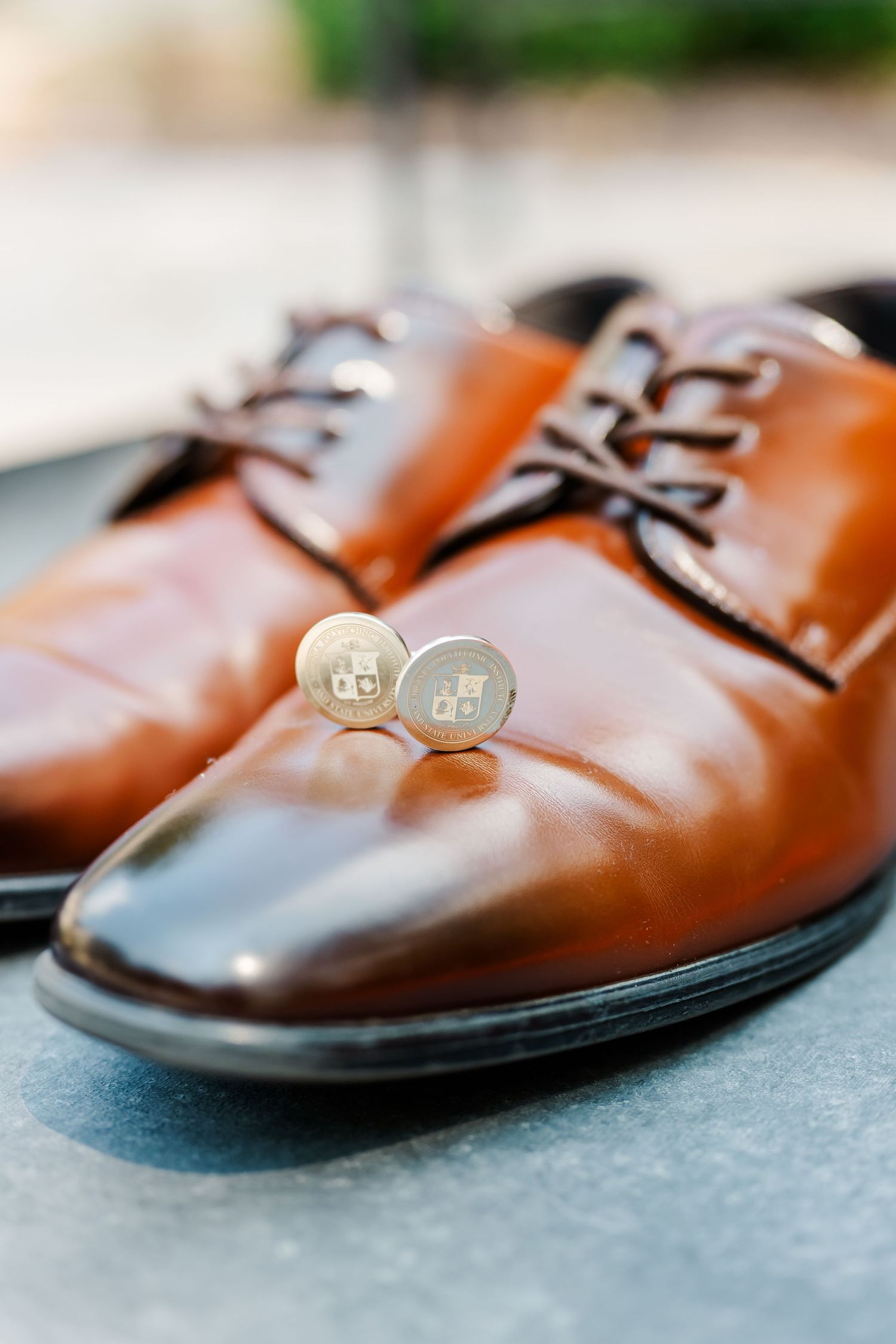 Silver monogram cufflinks rest in front of polished brown leather dress shoes on a gray surface.