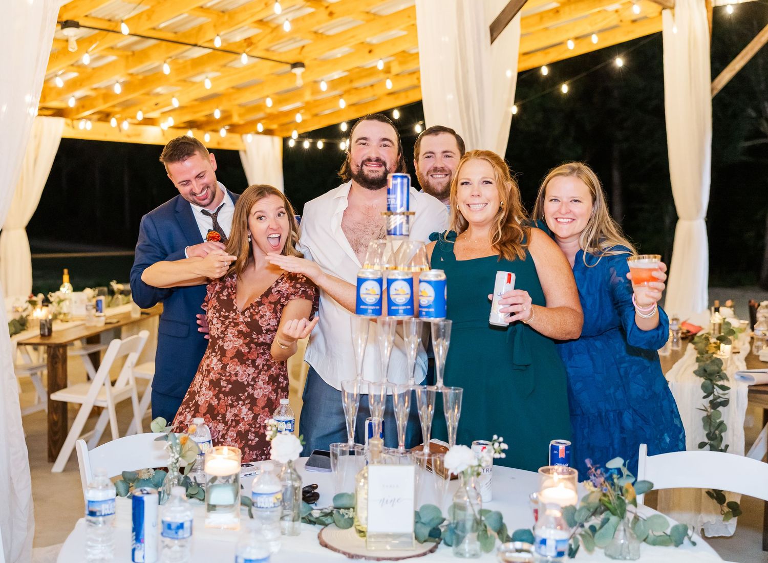 Group of six friends celebrating at a wedding reception, toasting with champagne glasses and drinks under string lights.